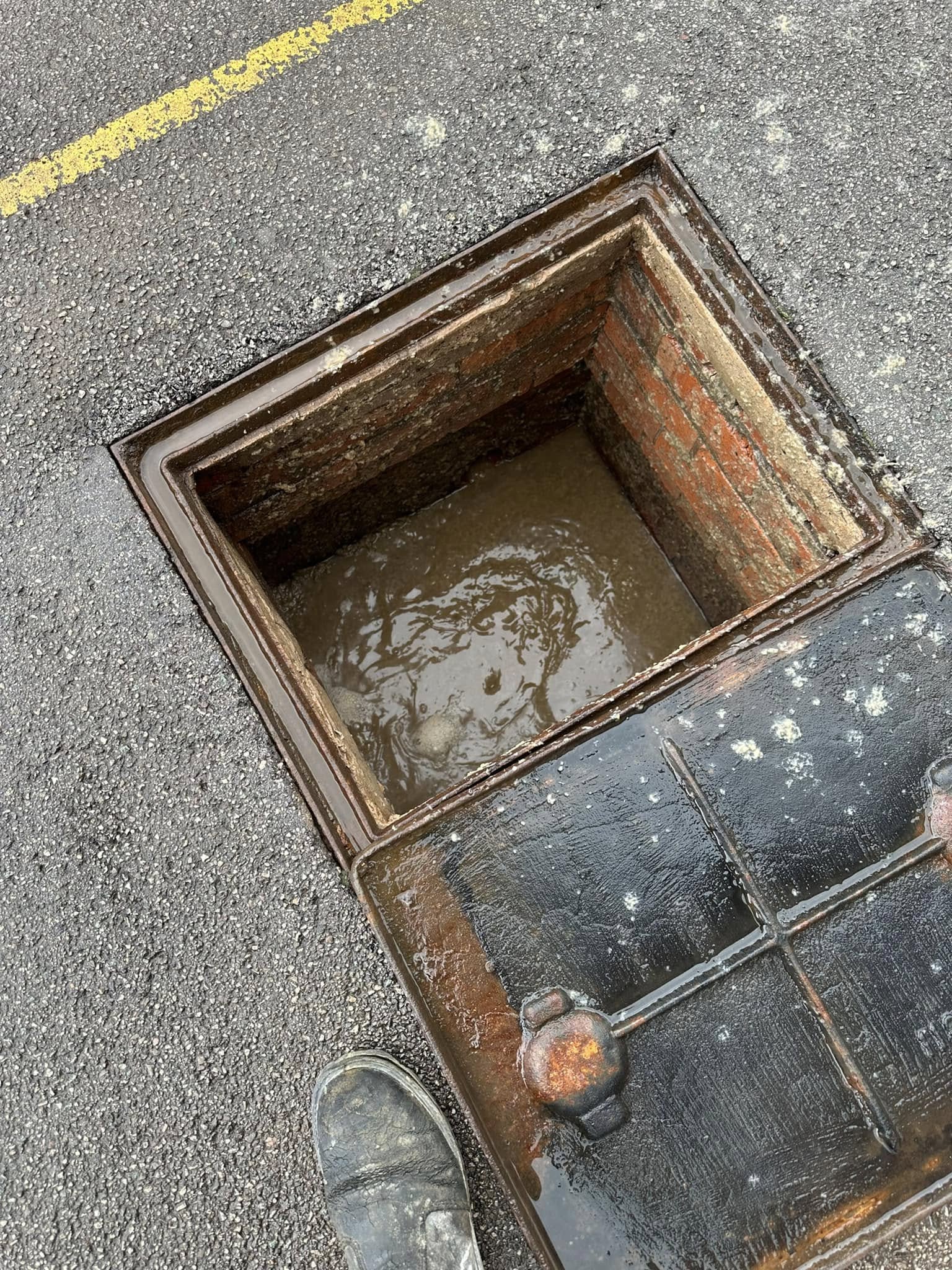 Close-up of a blocked drain, with murky wastewater visible in an open manhole on an asphalt surface.