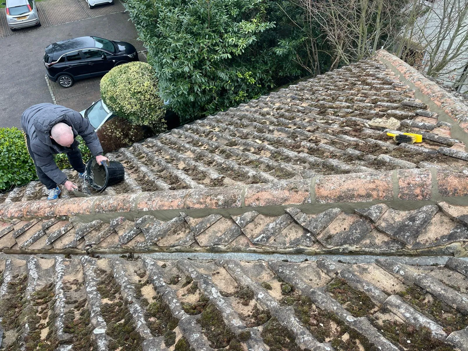 A roofer performing roof maintenance on an old tiled roof, applying fresh mortar to the ridges and valleys to secure tiles and prevent leaks. The roof is covered in moss, indicating a need for cleaning and general upkeep.