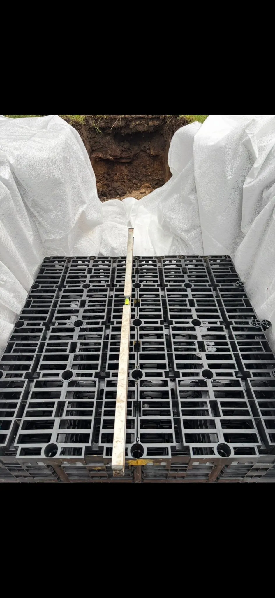 Modular drainage crates with a spirit level, inside an excavated pit lined with a white membrane, for a drainage system installation.