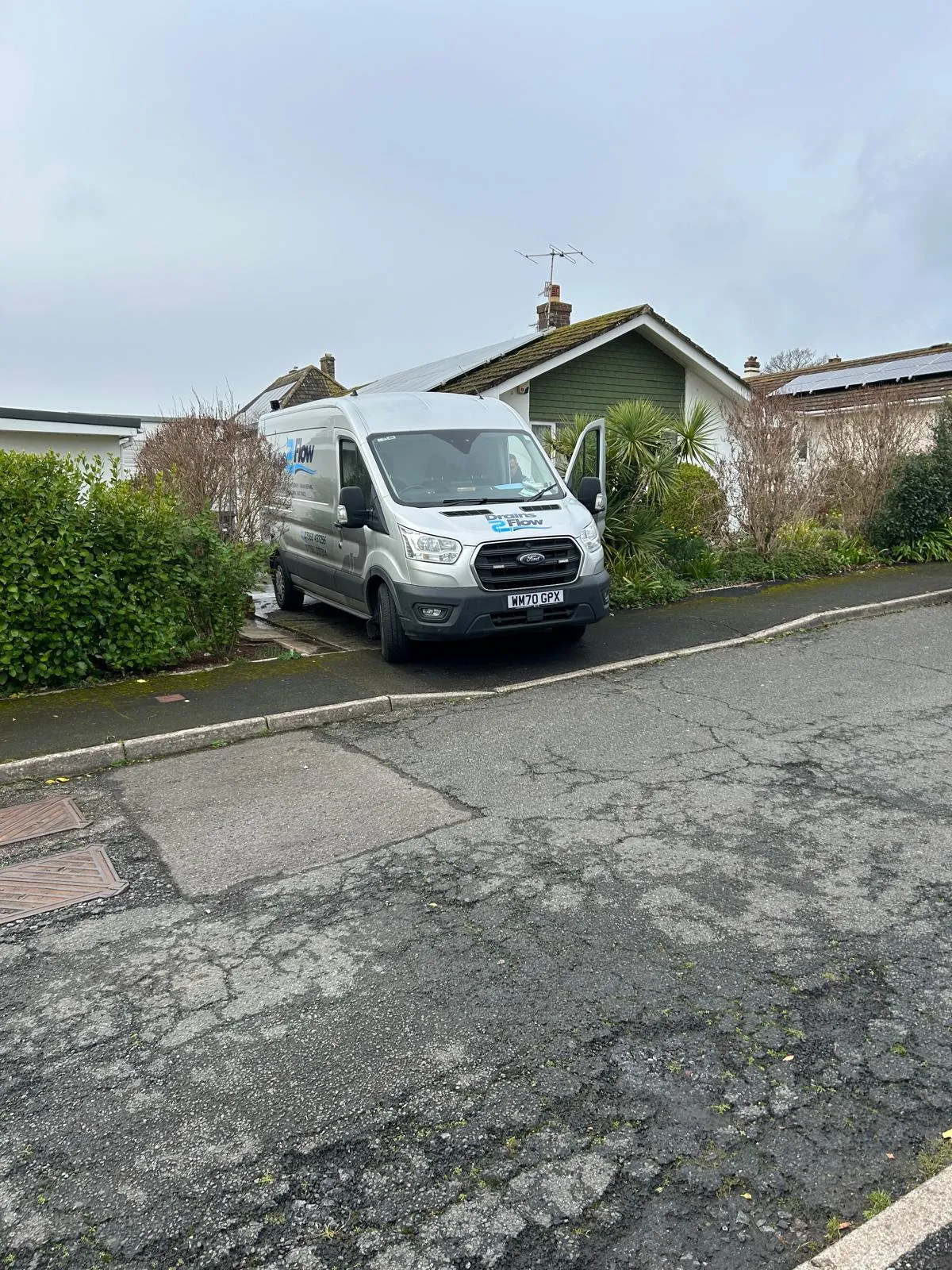 A silver Drains2Flow branded service van with the company logo on its side and door open, parked on a residential street in front of a house, indicating a drainage service call-out in Crediton or Devon.