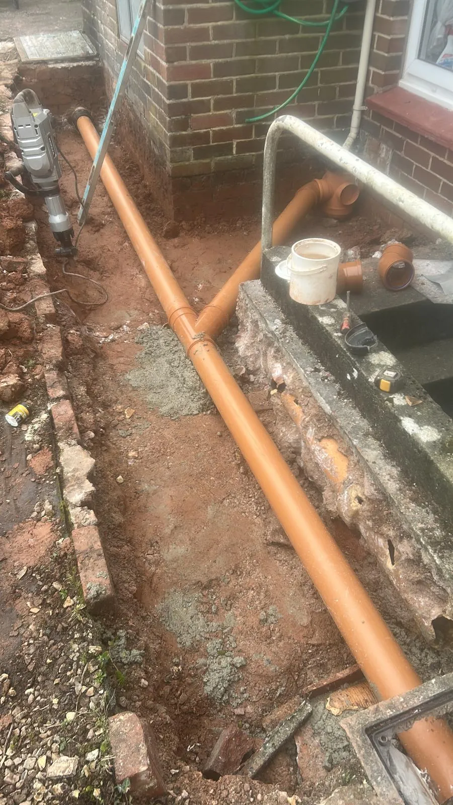 Installation of new orange drainage pipes in an excavated trench during groundworks near a building.