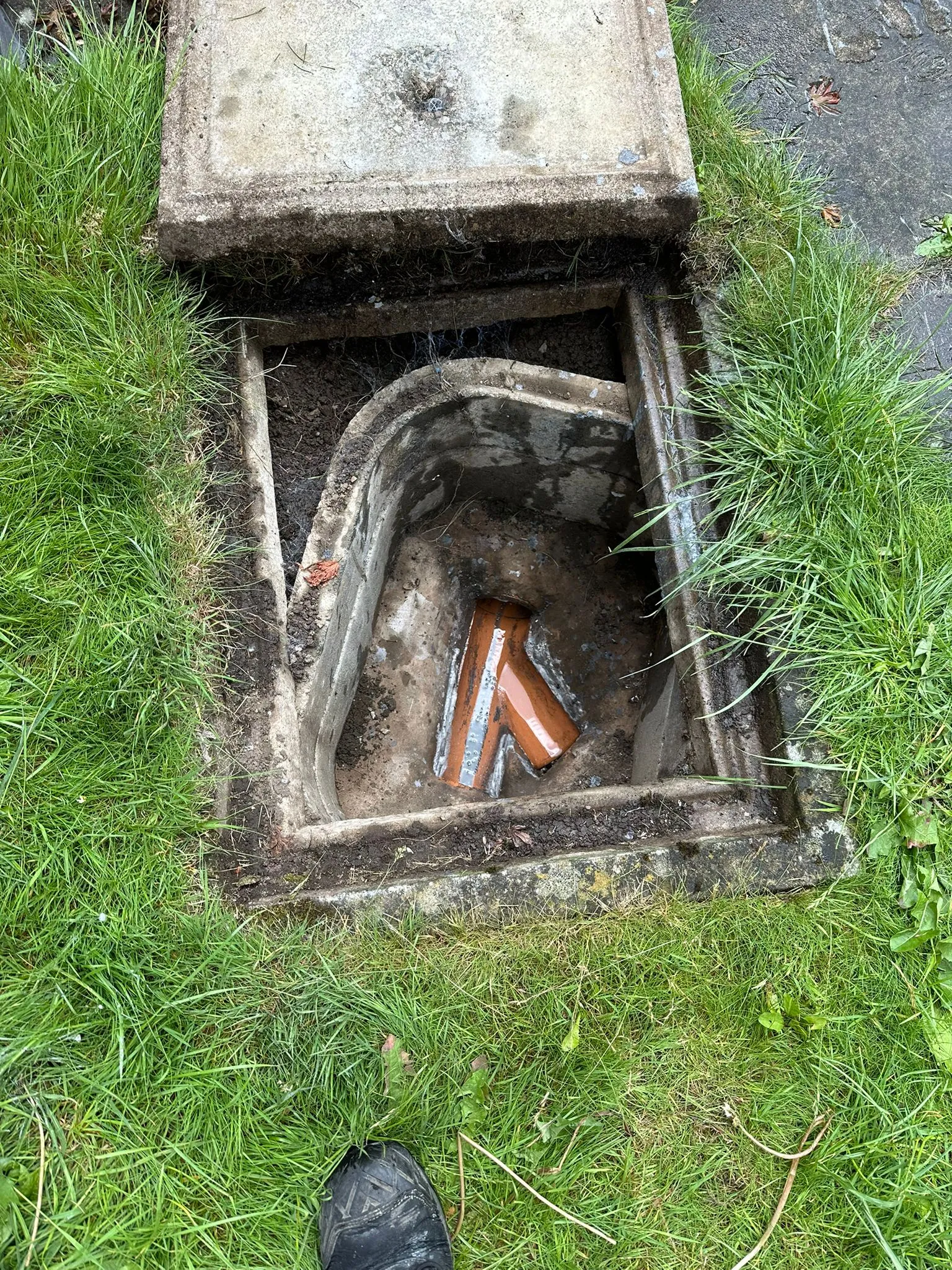 An open manhole cover revealing drainage pipes and the inspection chamber, set in a green grassy area. Used for drain surveys and unblocking services.