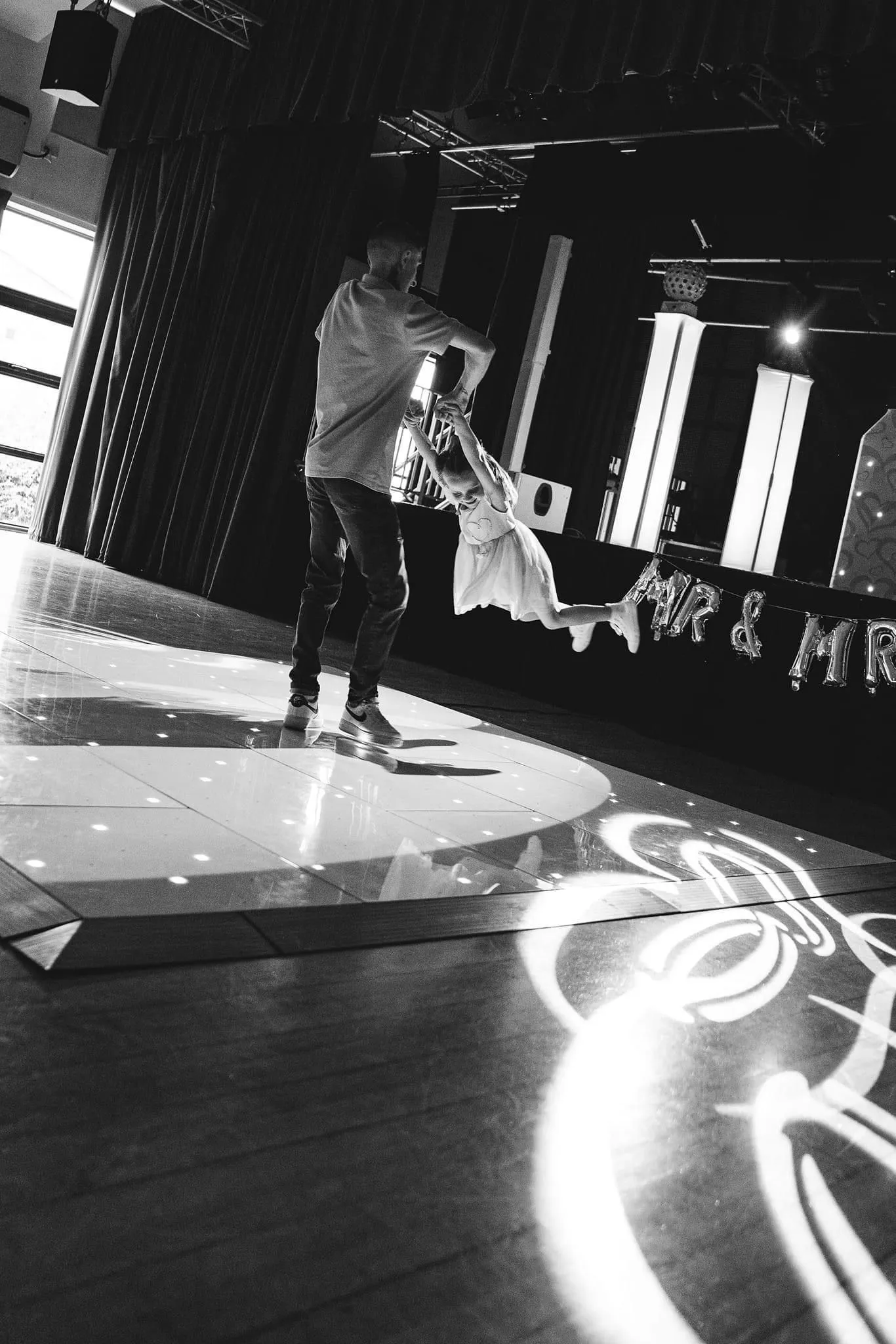 Adult swinging a child on a lighted LED dancefloor during a fun wedding reception, with 'Mr & Mr' balloons in the background.