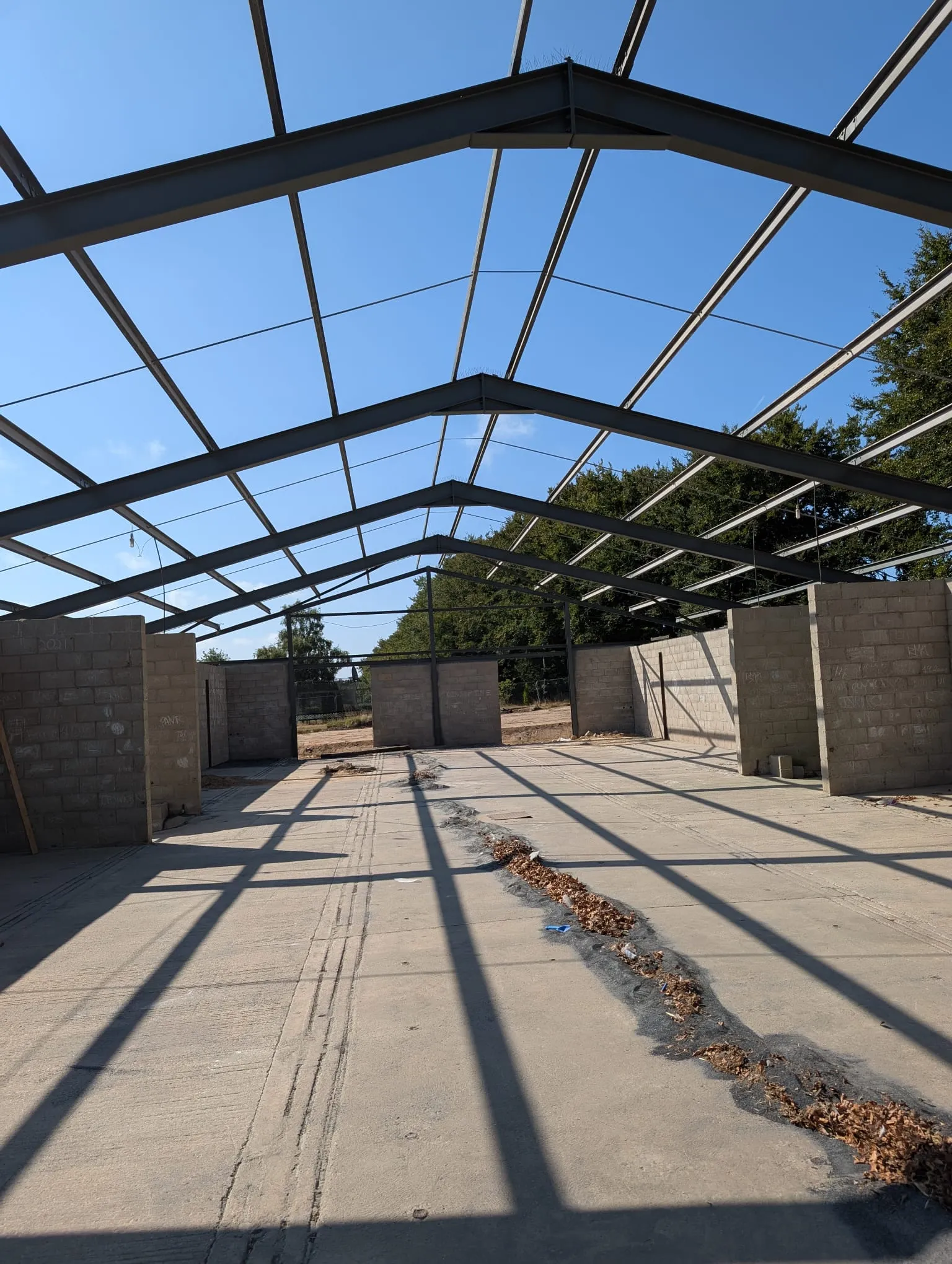 Interior view of a large, empty industrial building frame with steel girders and partial concrete block walls under a clear blue sky.