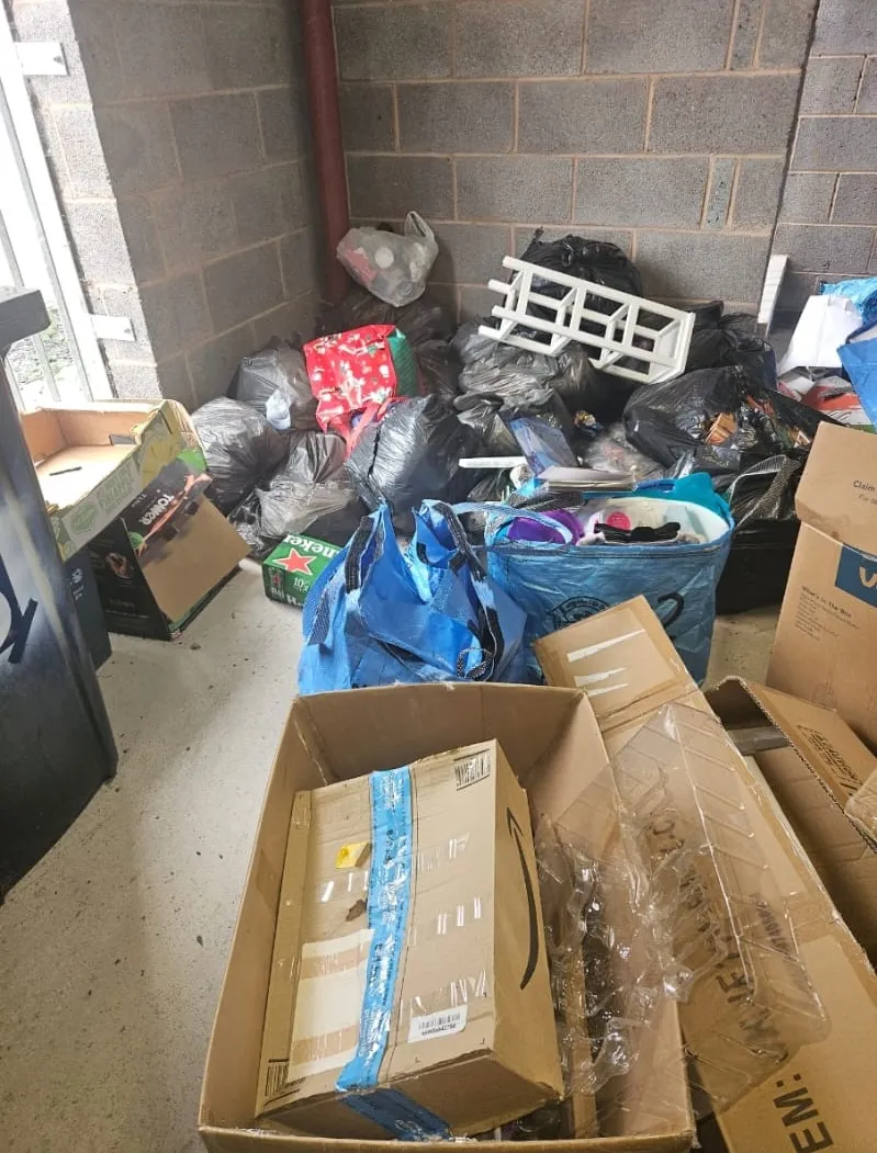 Cluttered garage corner filled with black bin bags, cardboard boxes, and miscellaneous rubbish awaiting collection.