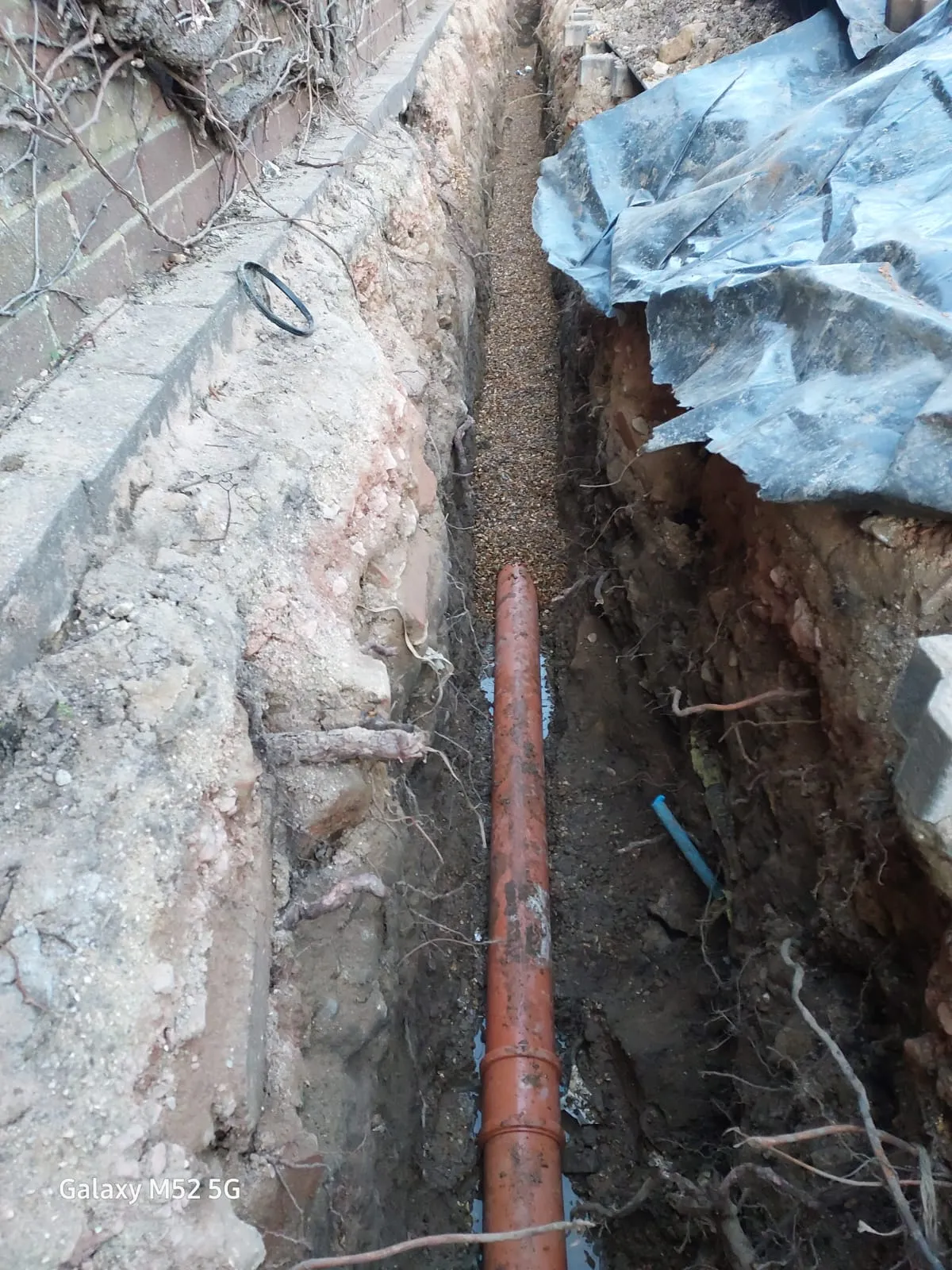 An orange drainage pipe placed in a deep trench with gravel, indicating new drain installation or repair work.