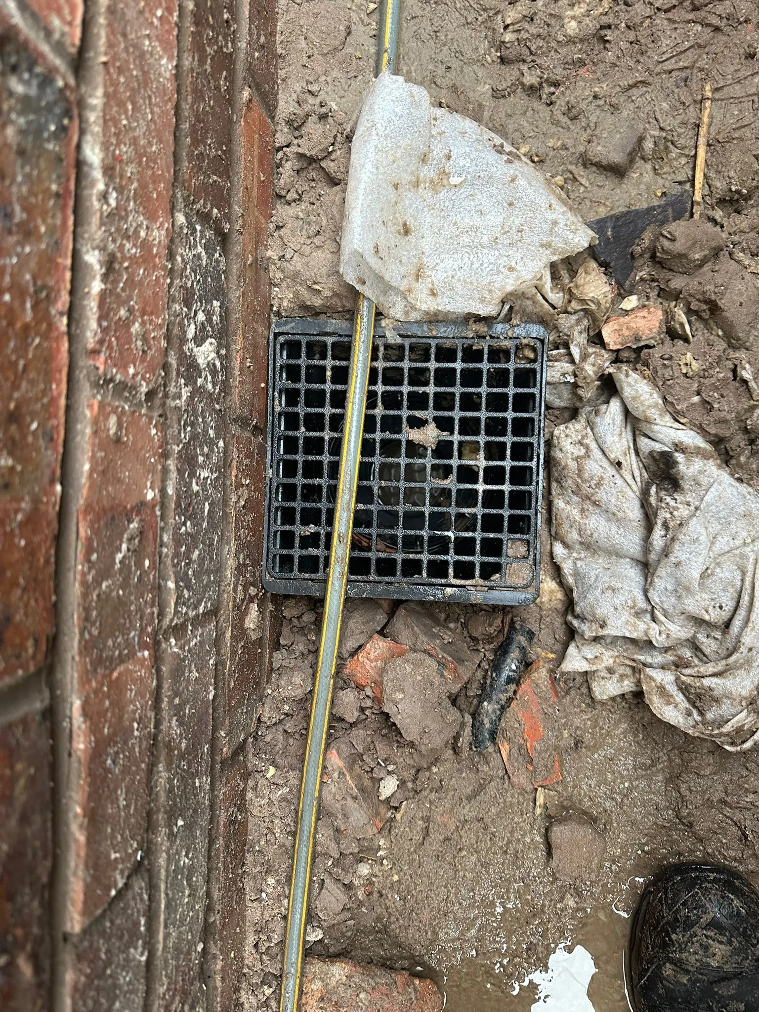 Outdoor gully drain surrounded by mud, debris, and a brick wall, with a hose running across the grate, indicating potential drainage work or a blockage.