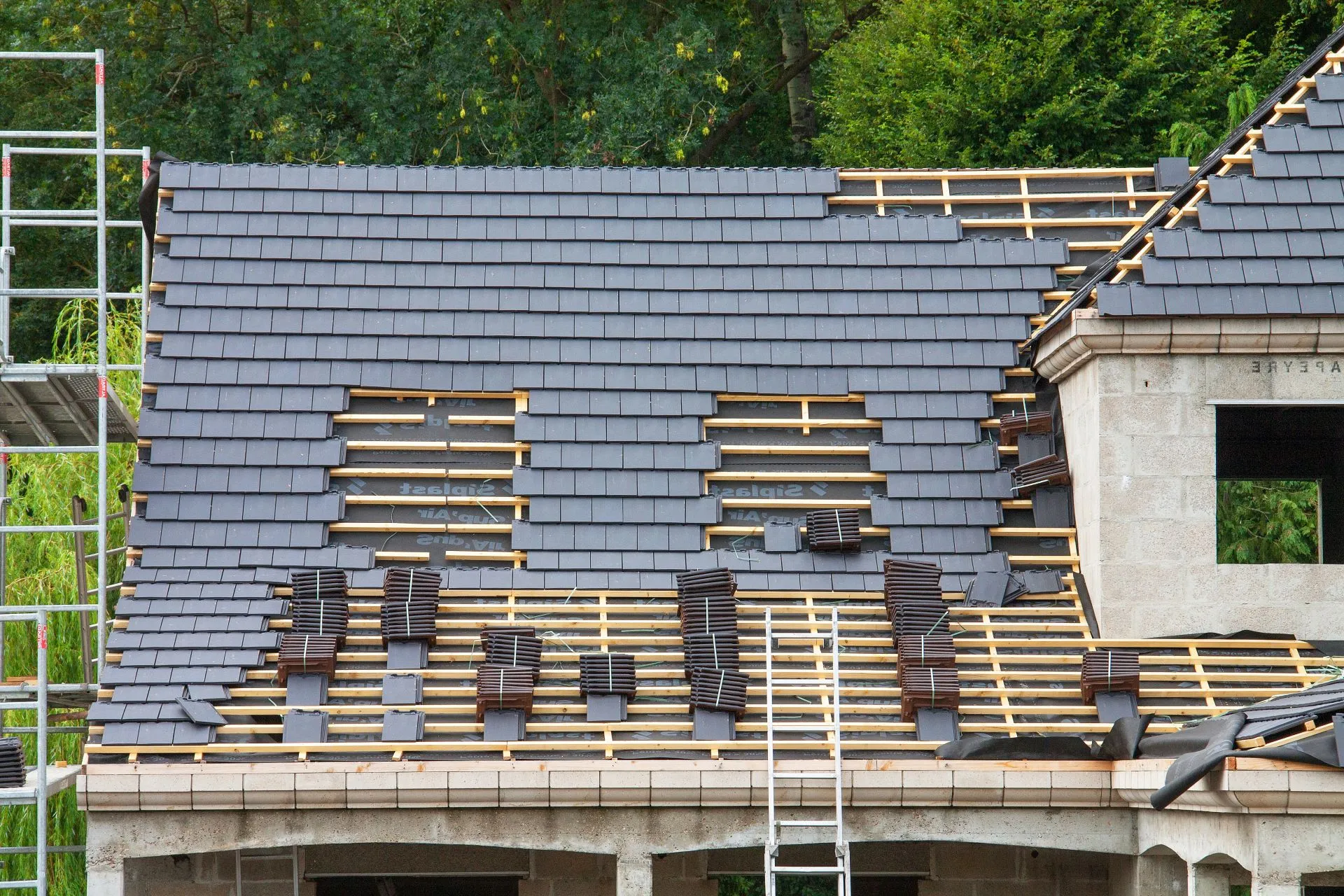 Installation of new dark grey roof tiles on a pitched roof, with visible roof structure and scaffolding during construction.