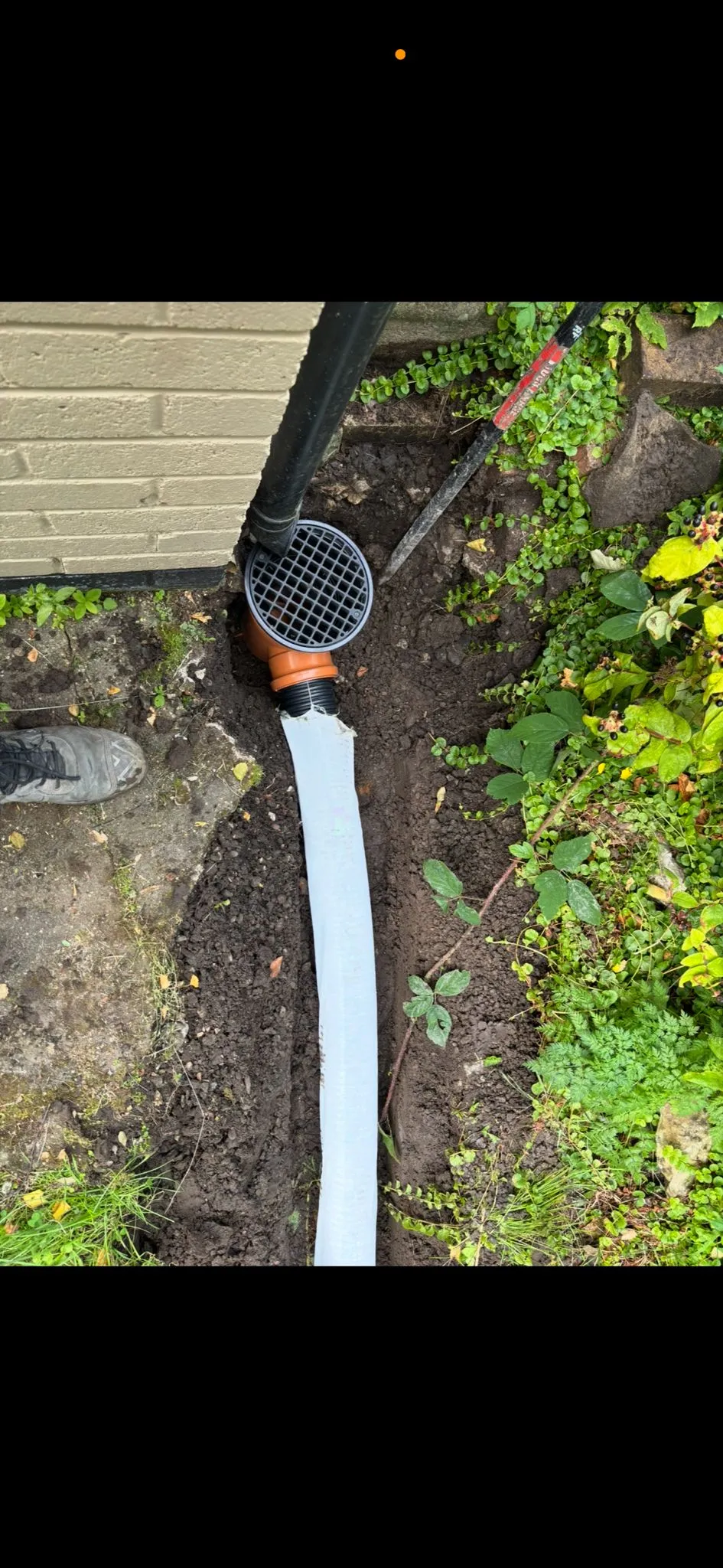 Newly installed gully and drainage pipe in an excavated trench, connecting to a building's downspout.