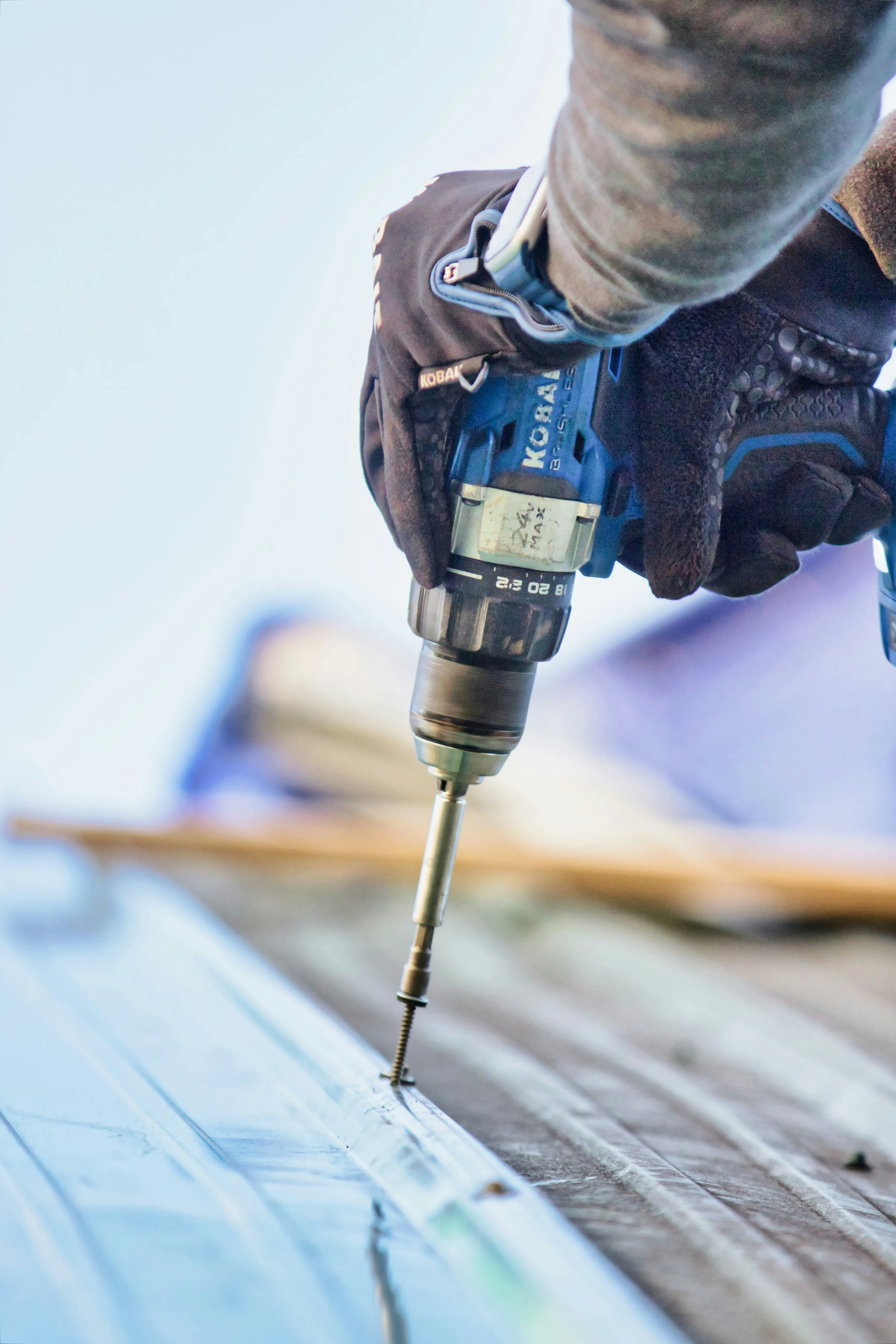 A roofer's gloved hand using a power drill to install a screw into a new metal roof.