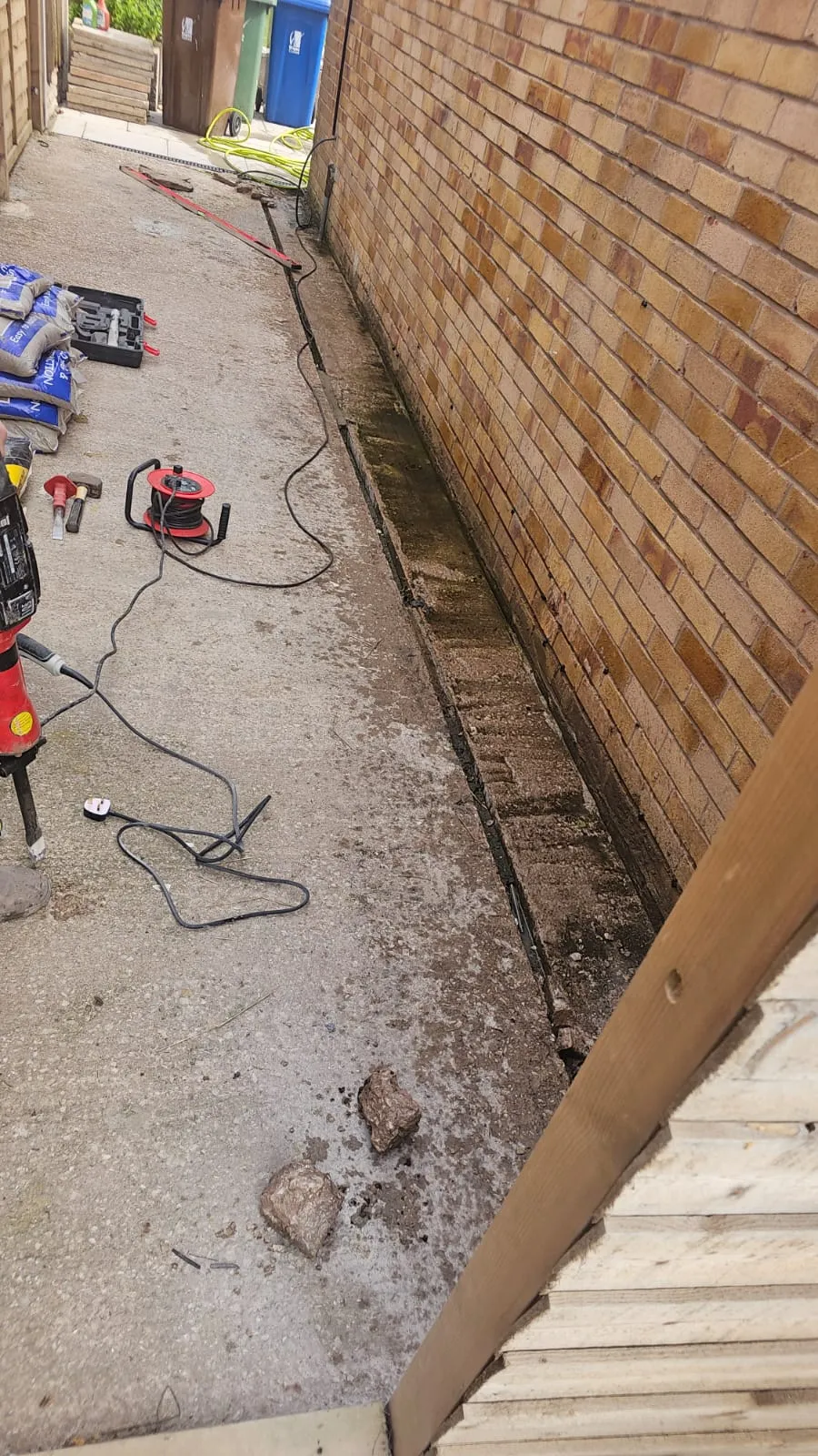 Drain excavation site showing a dug trench along a brick wall with various tools and materials.