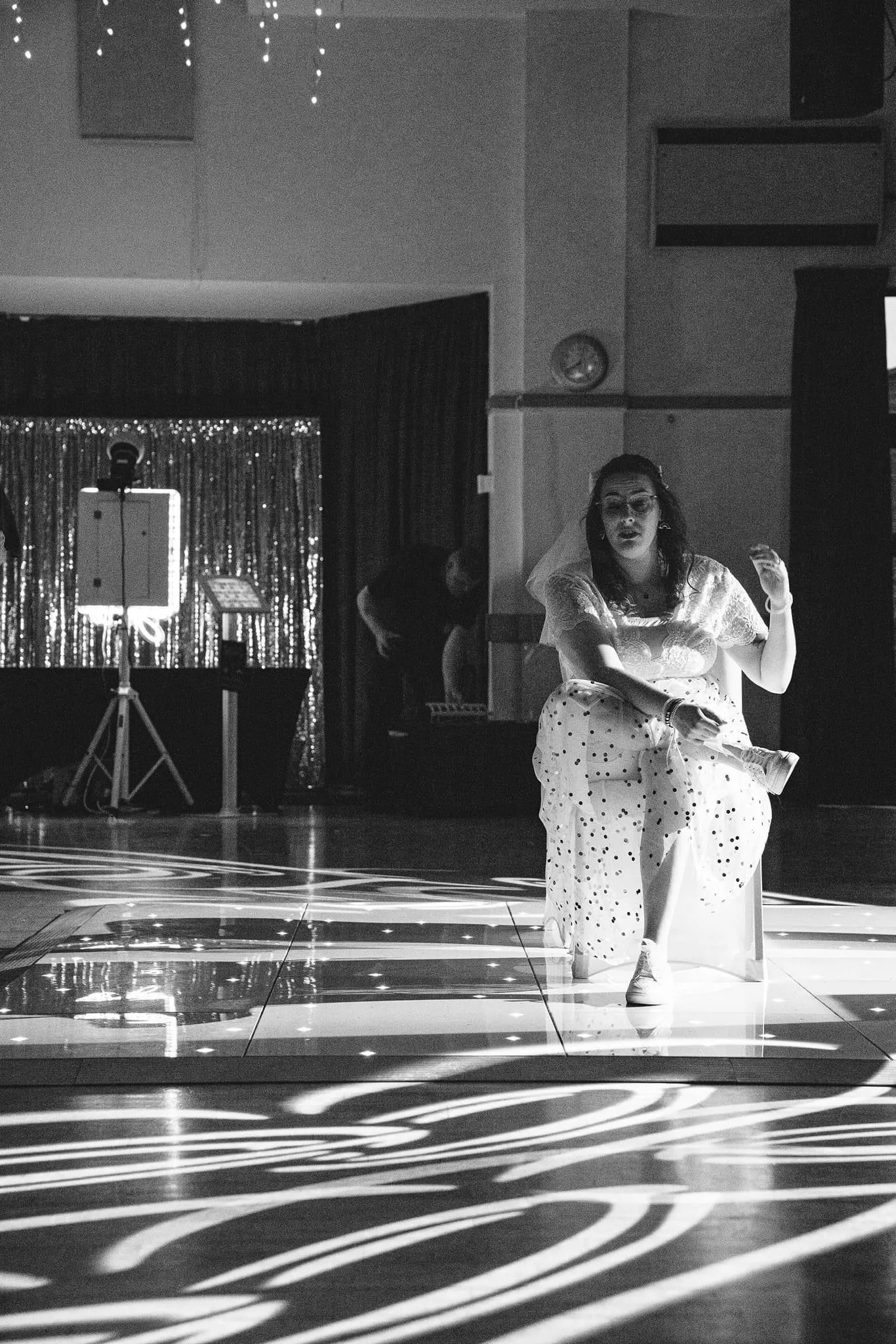 Black and white photo of a woman in a dress sitting on a reflective dance floor with DJ equipment and a sparkling backdrop in the background, possibly at a wedding reception.