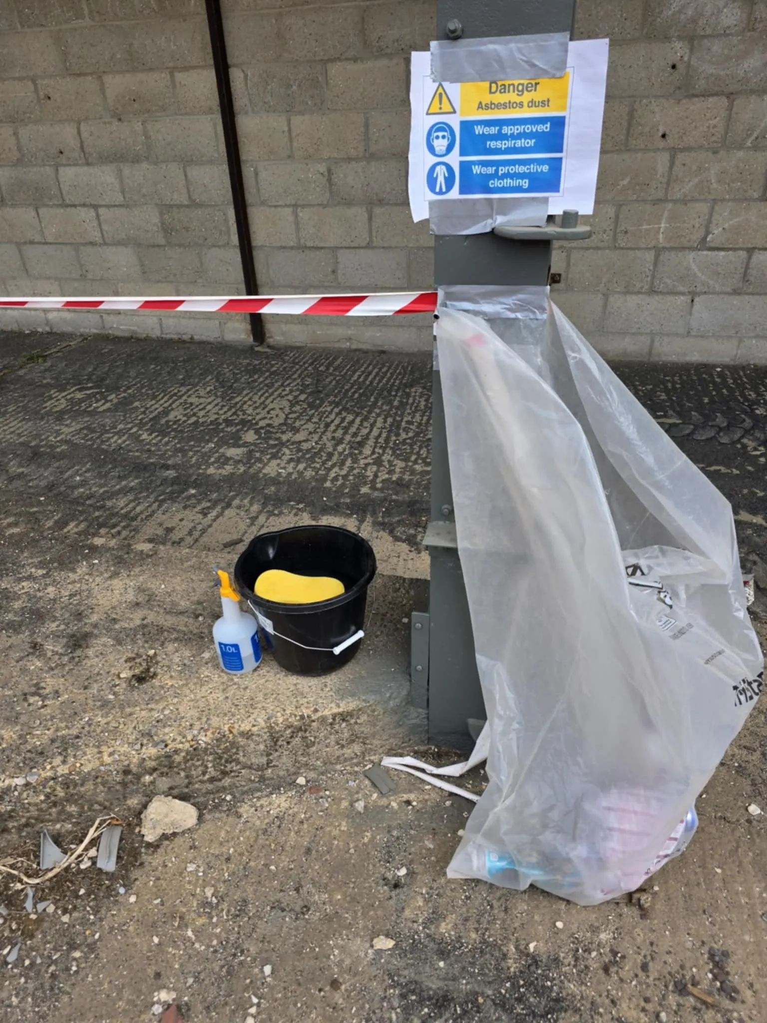 An asbestos removal work area showing a 'Danger Asbestos dust' sign, mandatory respirator and protective clothing symbols, barrier tape, a black bucket with a yellow sponge, and a spray bottle.
