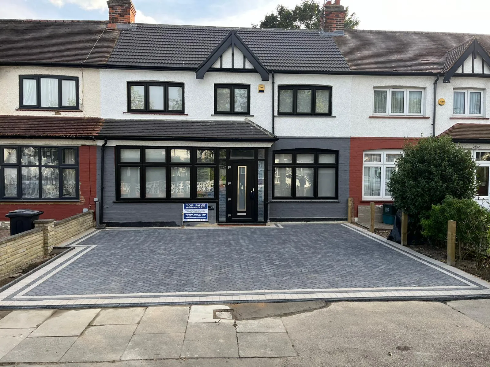A modern grey block paving driveway with a light border, laid in a herringbone pattern, in front of a renovated terraced house.