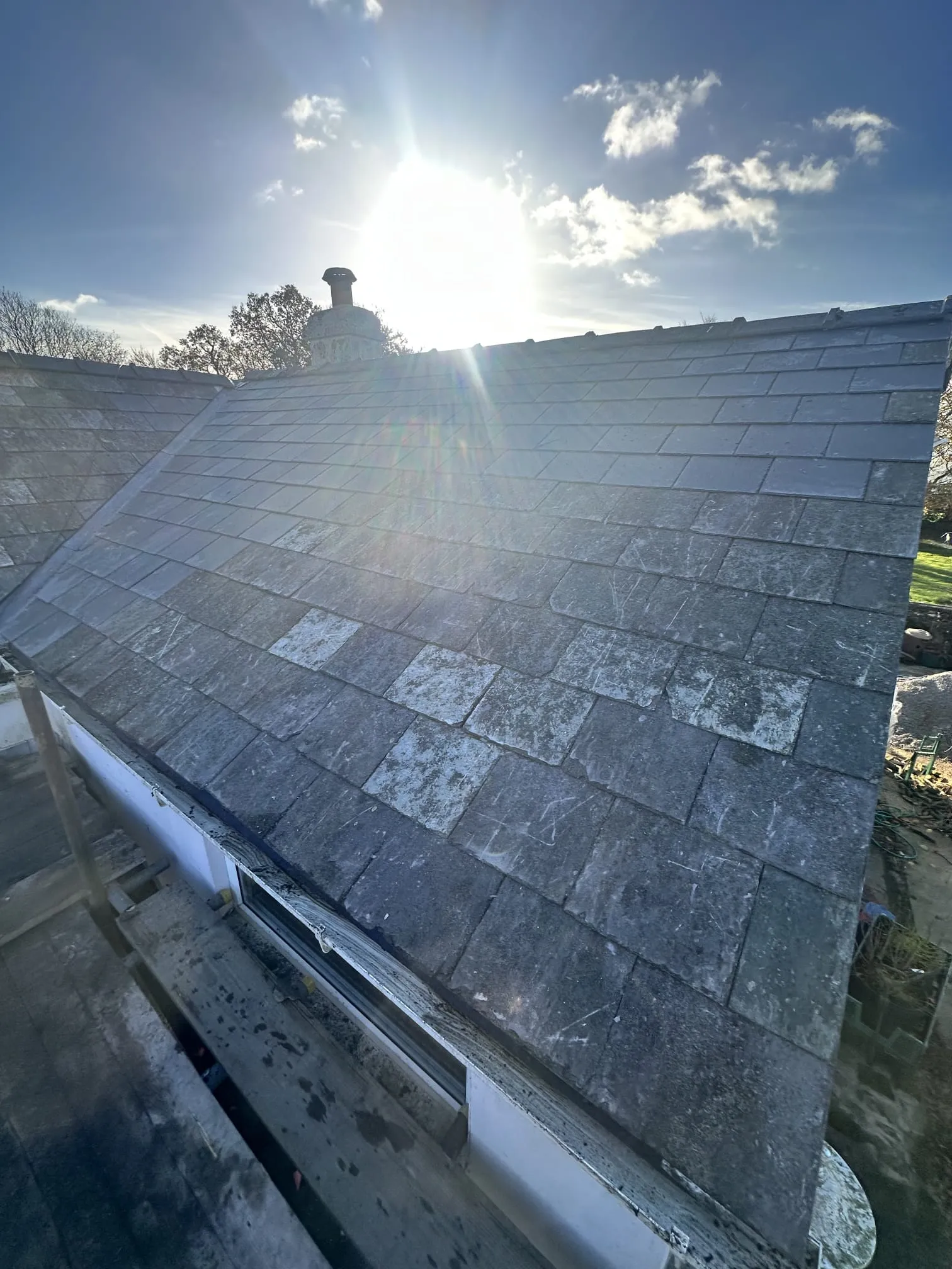 A pitched slate roof on a sunny day, with some tiles appearing worn or discoloured, and a chimney stack in the background, hinting at roofing repair or cleaning work.
