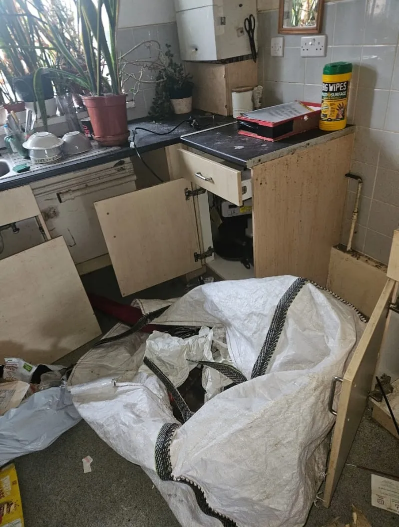 A heavily cluttered and untidy kitchen with a large bag of waste, depicting a challenging house clearance scenario or hoarder clean-up.