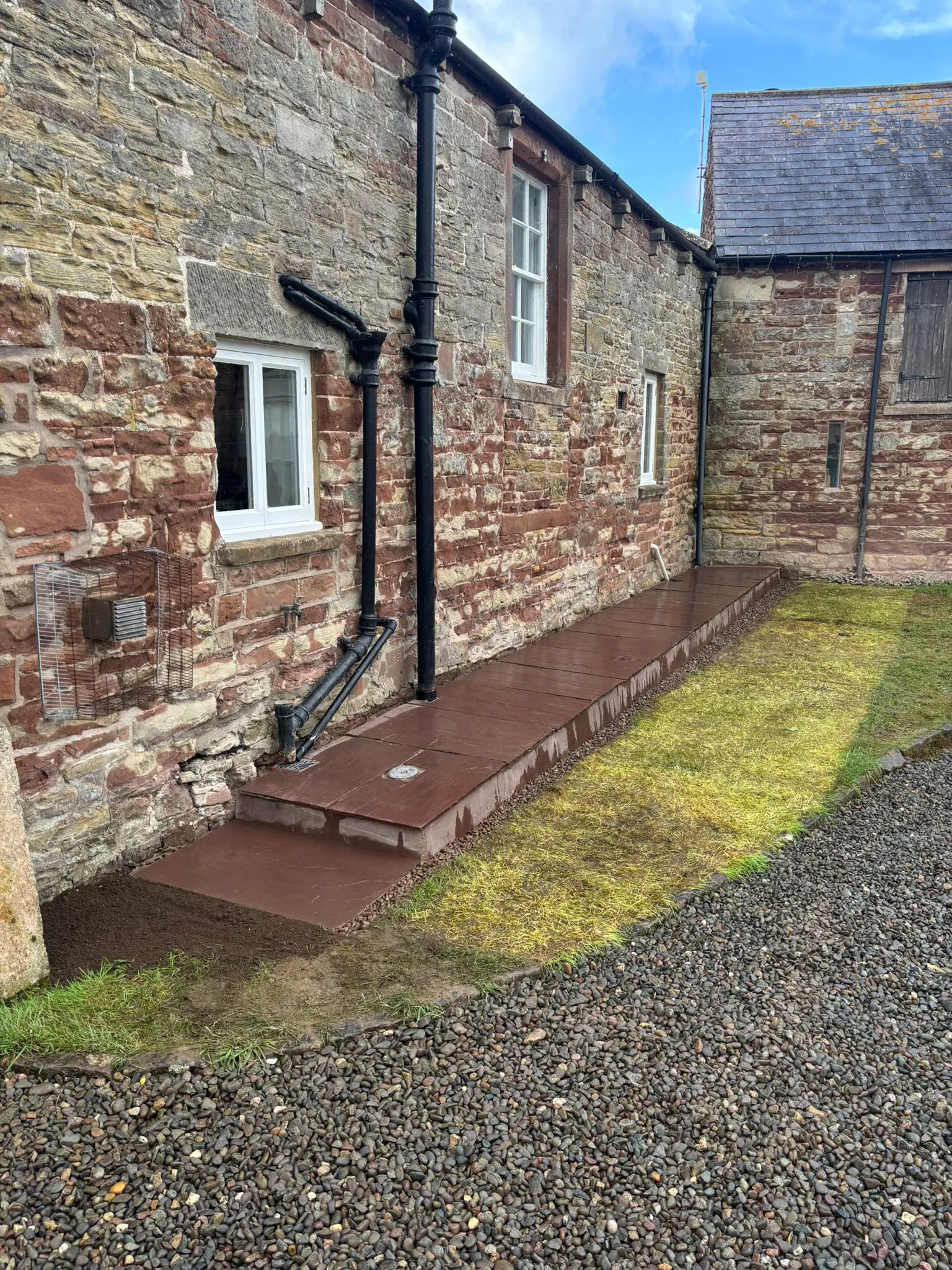 Newly laid paved pathway and garden landscaping next to an old stone building with drainage.