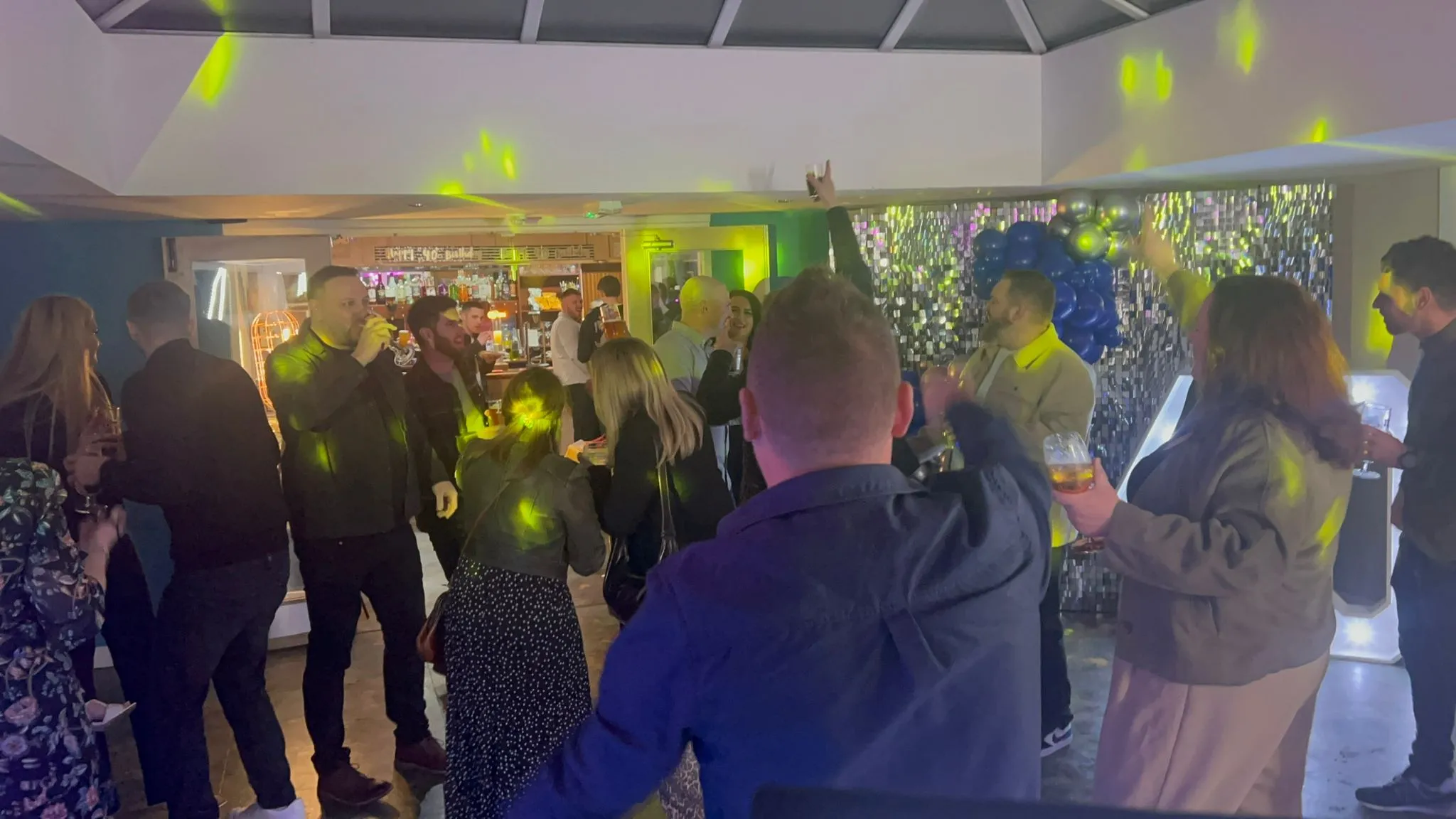 A group of adults dancing and socialising at a party, illuminated by yellow disco lights, with drinks in hand and a bar visible in the background.
