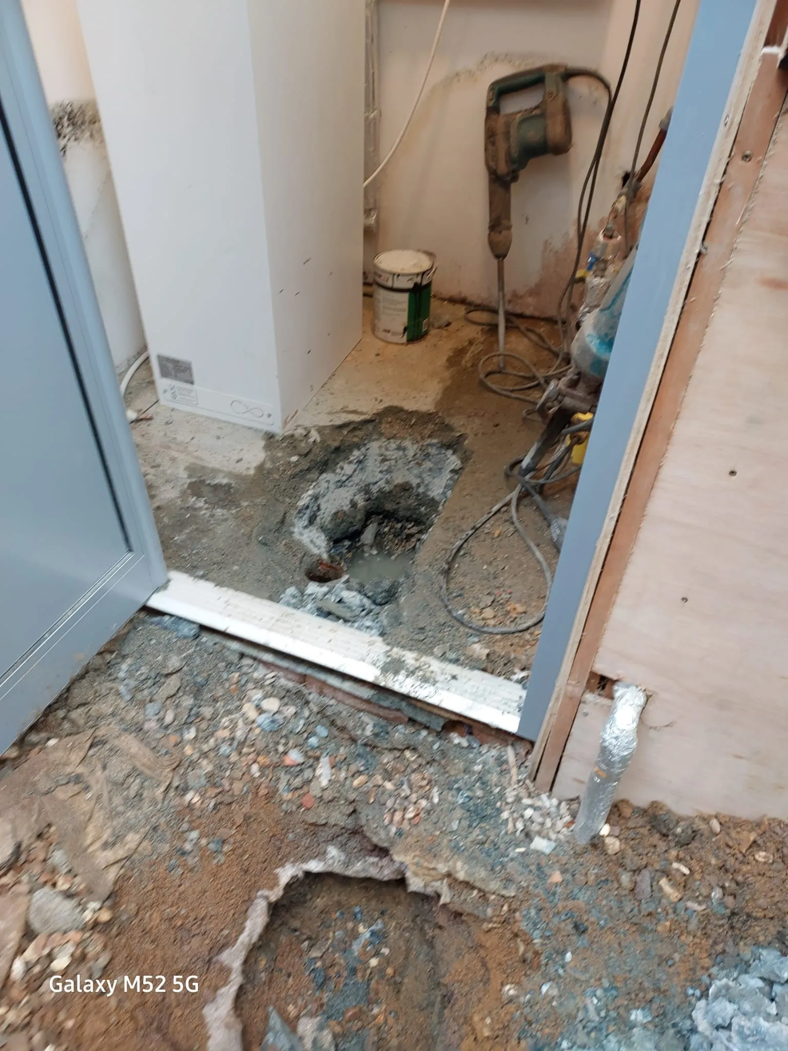 An interior shot of a room with a section of the concrete floor excavated, revealing a drainage pipe and soil, with a jackhammer and other tools on the ground.