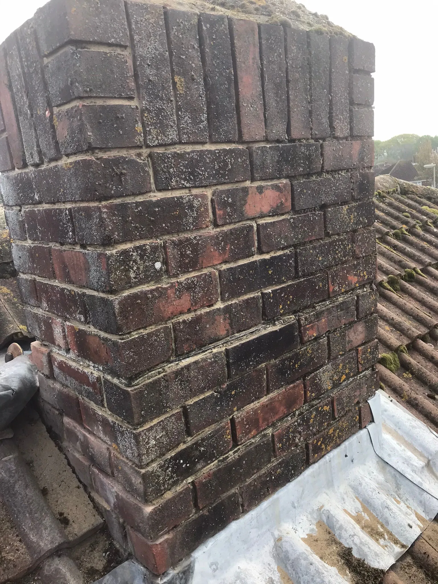 A weathered brick chimney stack on a tiled roof with lead flashing, showing signs of moss and lichen growth.