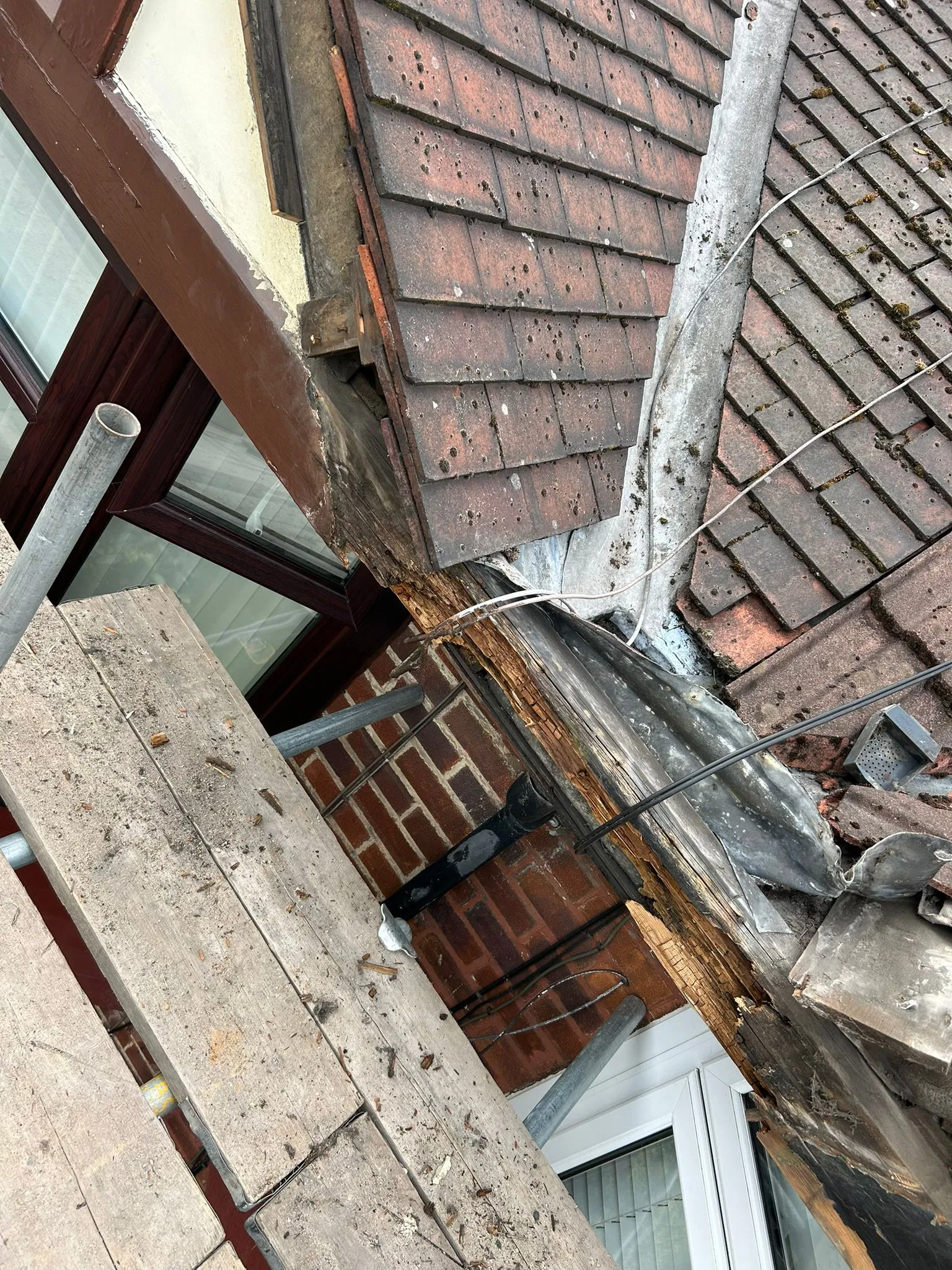 A close-up of a severely damaged roof with exposed rotten timber, red clay tiles, and old lead flashing, viewed from scaffolding.