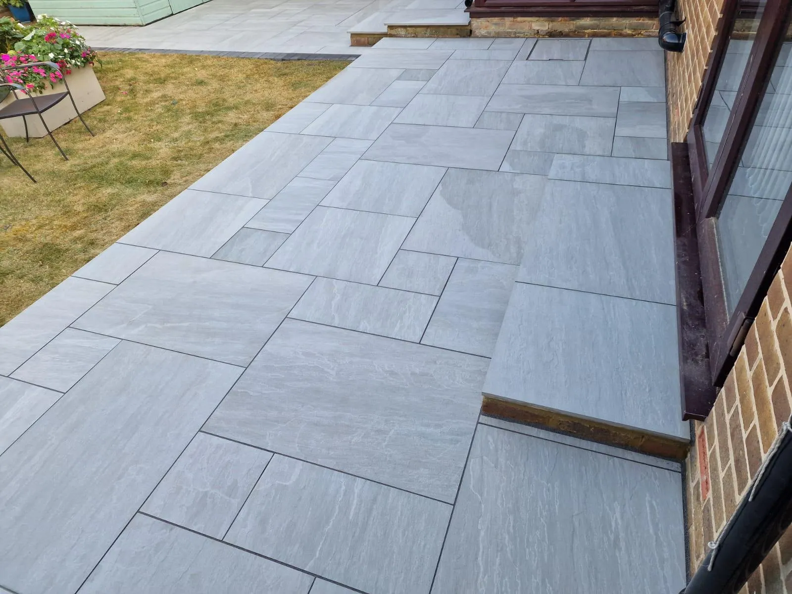 A contemporary grey patio with large format paving slabs next to a house and a small garden area, showing a fresh installation.
