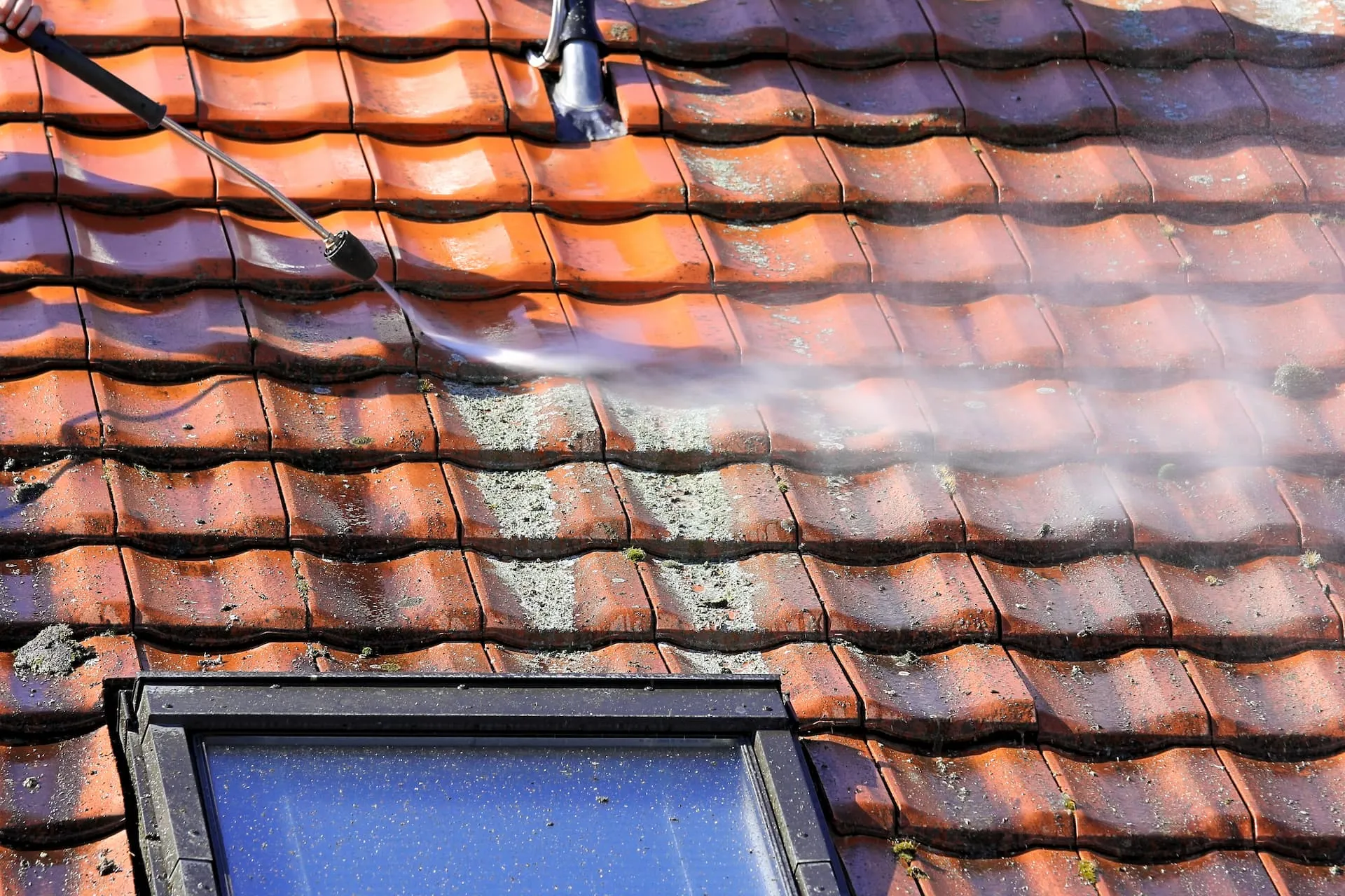 A person using a pressure washer to clean moss and grime from terracotta roof tiles, revealing cleaner sections, with a roof window in the foreground.