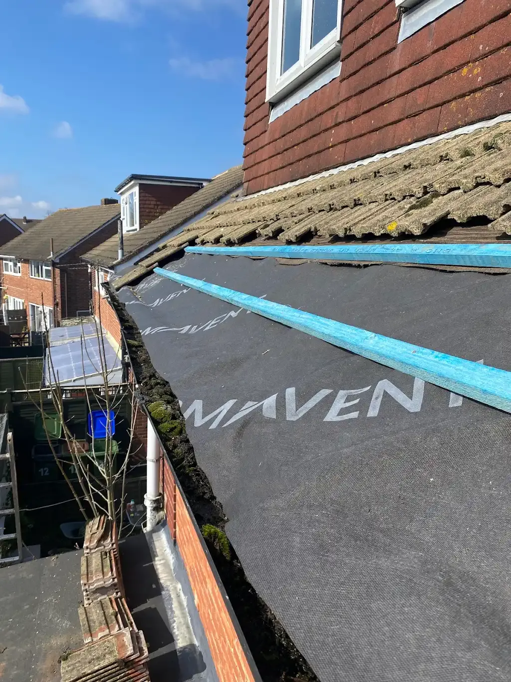 Residential roof replacement/repair work in progress, showing exposed underlayment, roof battens, and a blocked gutter filled with moss and debris.