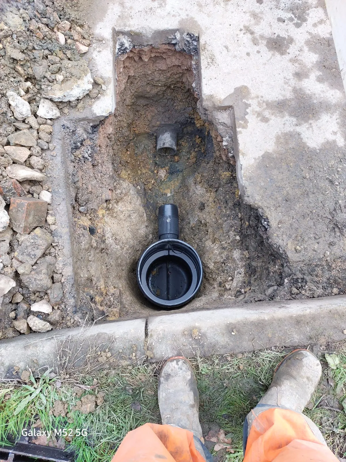 Top-down view of an excavated trench with a newly installed black non-return valve and an older grey pipe, surrounded by soil, rubble, and concrete.