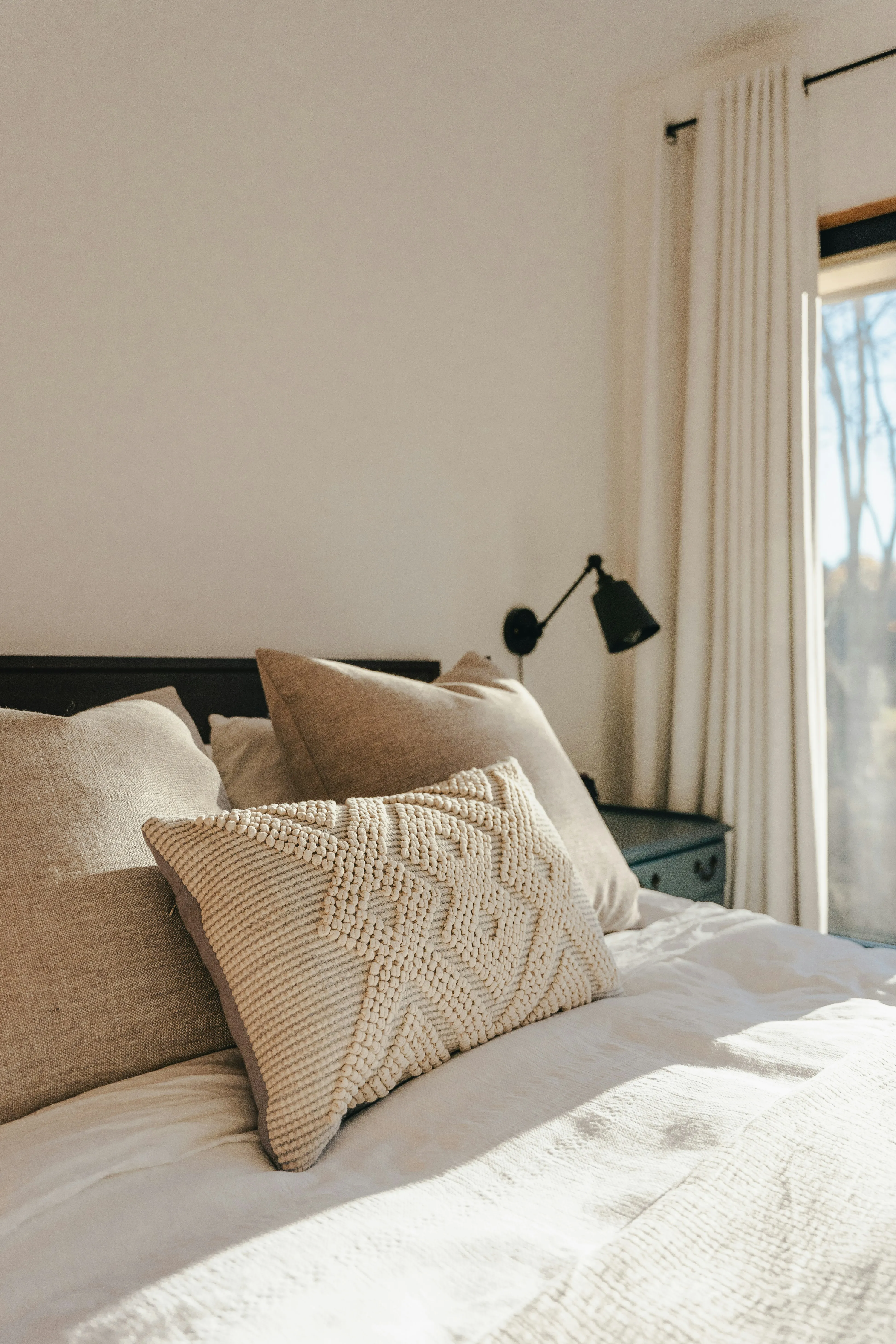 A comfortable bed with neutral-coloured pillows and bedding, bathed in sunlight from a nearby window.