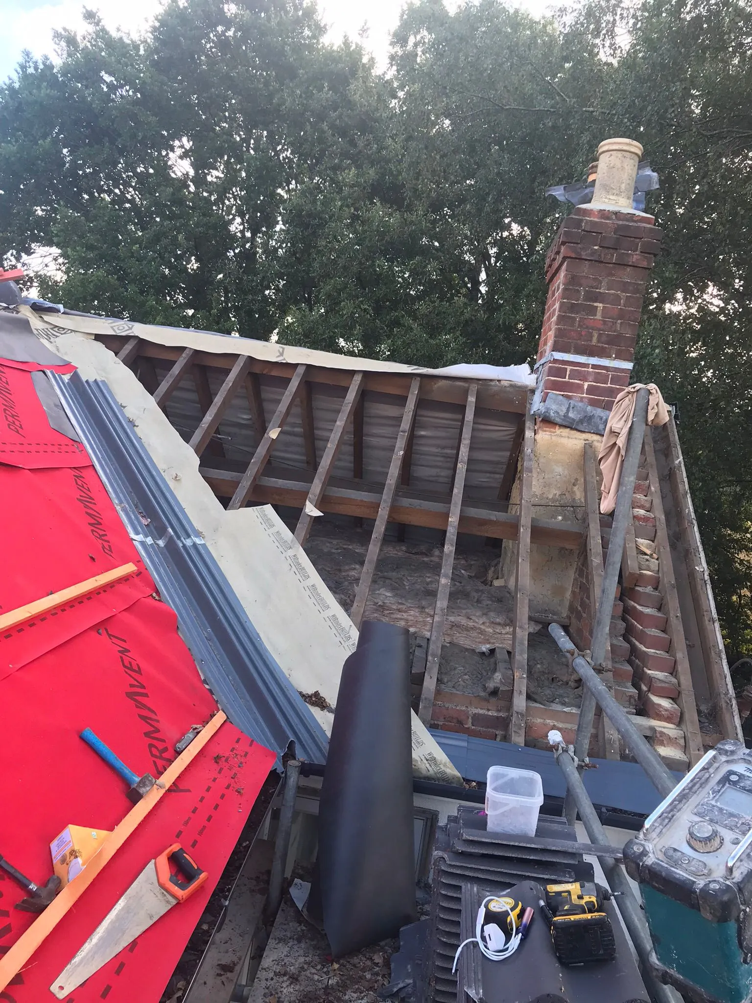 Pitched roof repair and renovation in progress, showing exposed wooden rafters, red roofing membrane, a brick chimney, and construction tools.