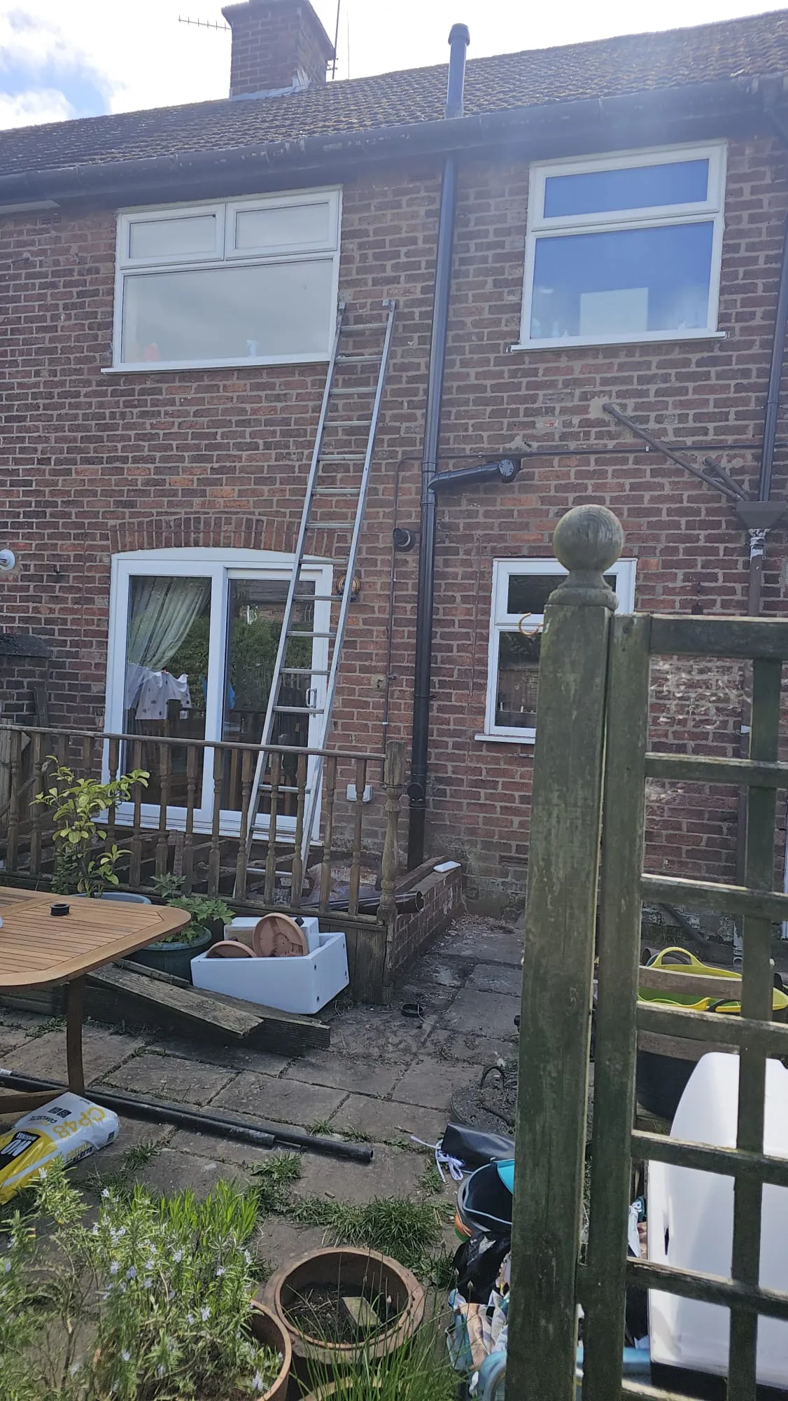 A residential brick house with a ladder leaning against it, showing external drainage pipes and a patio.