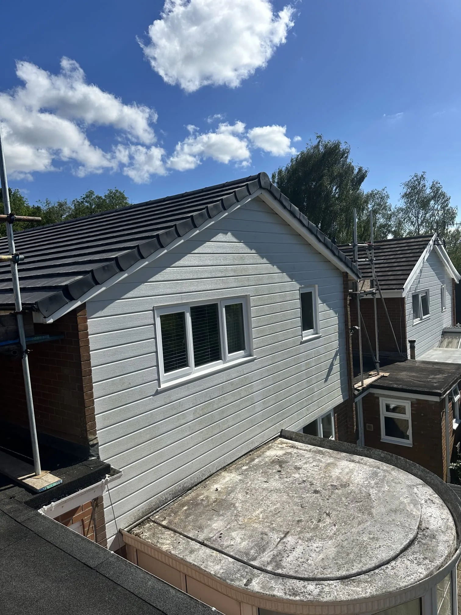 Residential homes with new dark grey tiled roofs, light grey cladding, and visible scaffolding. An old, worn flat roof is in the mid-ground, and a newer black flat roof is in the foreground.