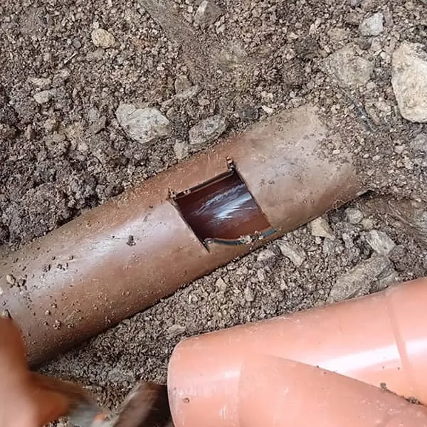 A broken brown drainage pipe exposed in the ground with a large rectangular hole, suggesting severe damage.