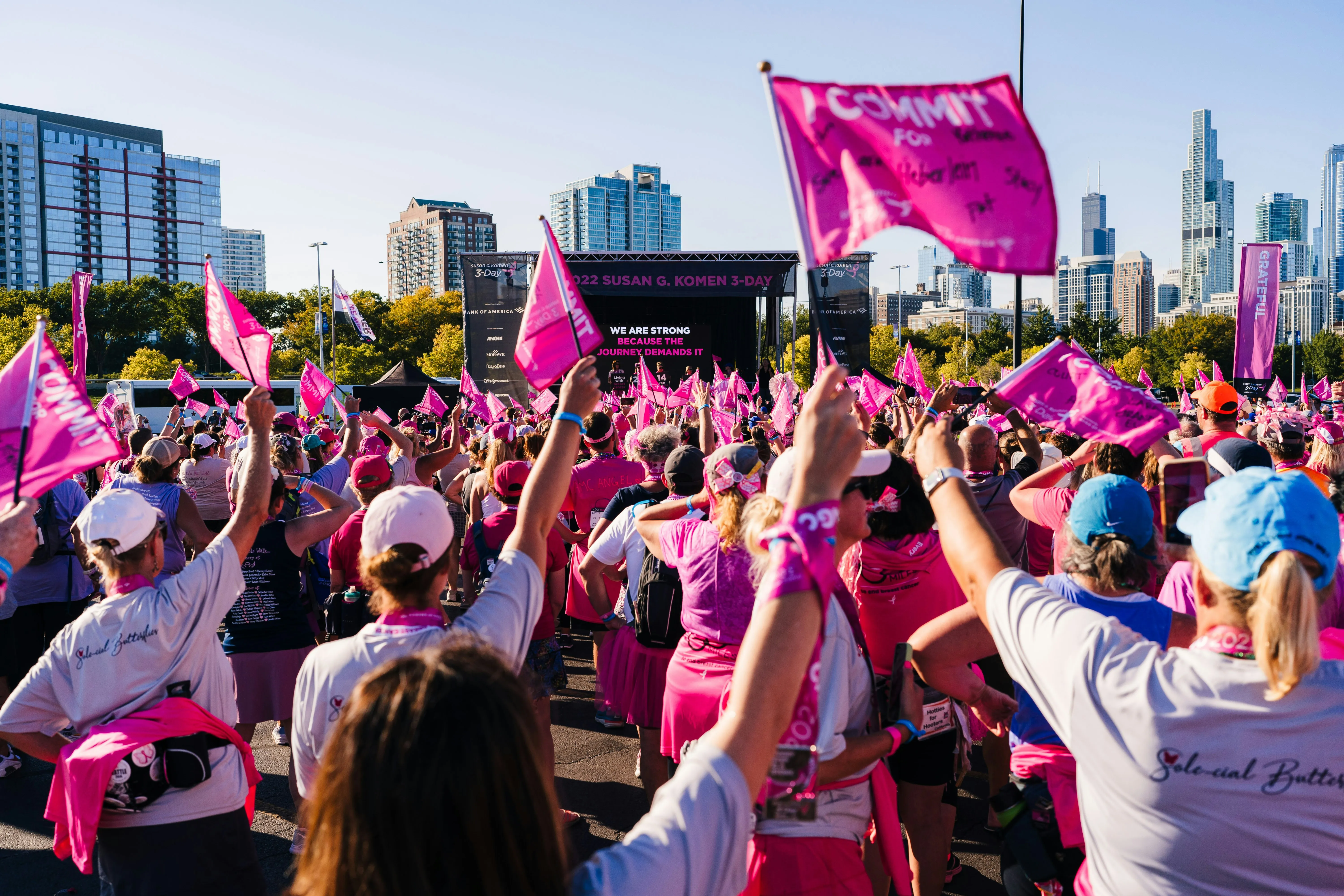 Participants at the 2022 Susan G. Komen 3-Day walk raise pink flags in celebration, with city skyscrapers and an event stage visible behind them.