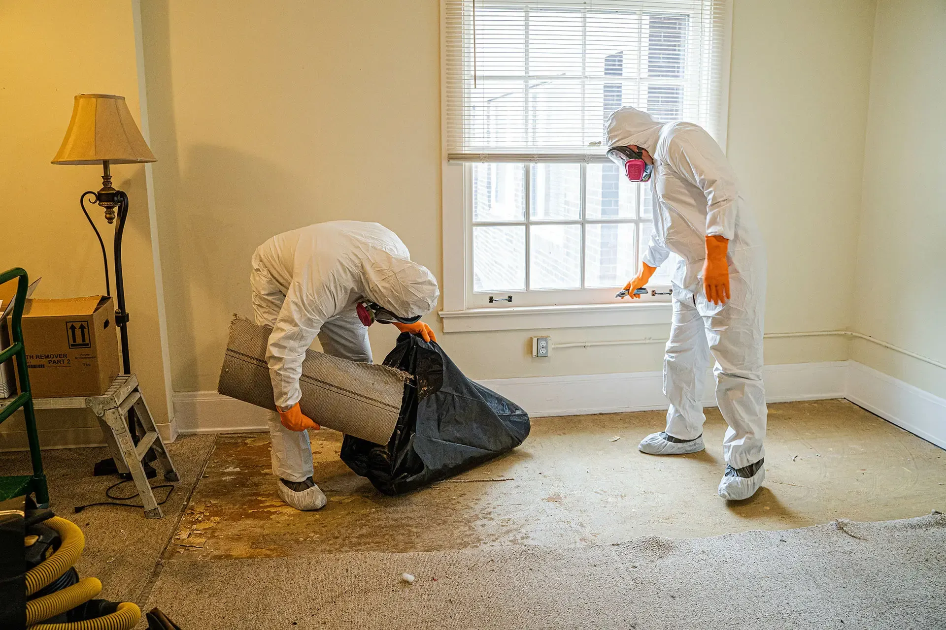 Two asbestos removal experts in hazmat suits safely removing old flooring materials and adhesive from a domestic room.
