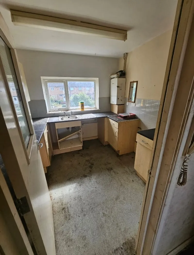 A dirty and unkempt kitchen in an empty property, showing old wooden cabinets, a sink, and a stained floor in need of clearance and cleaning.