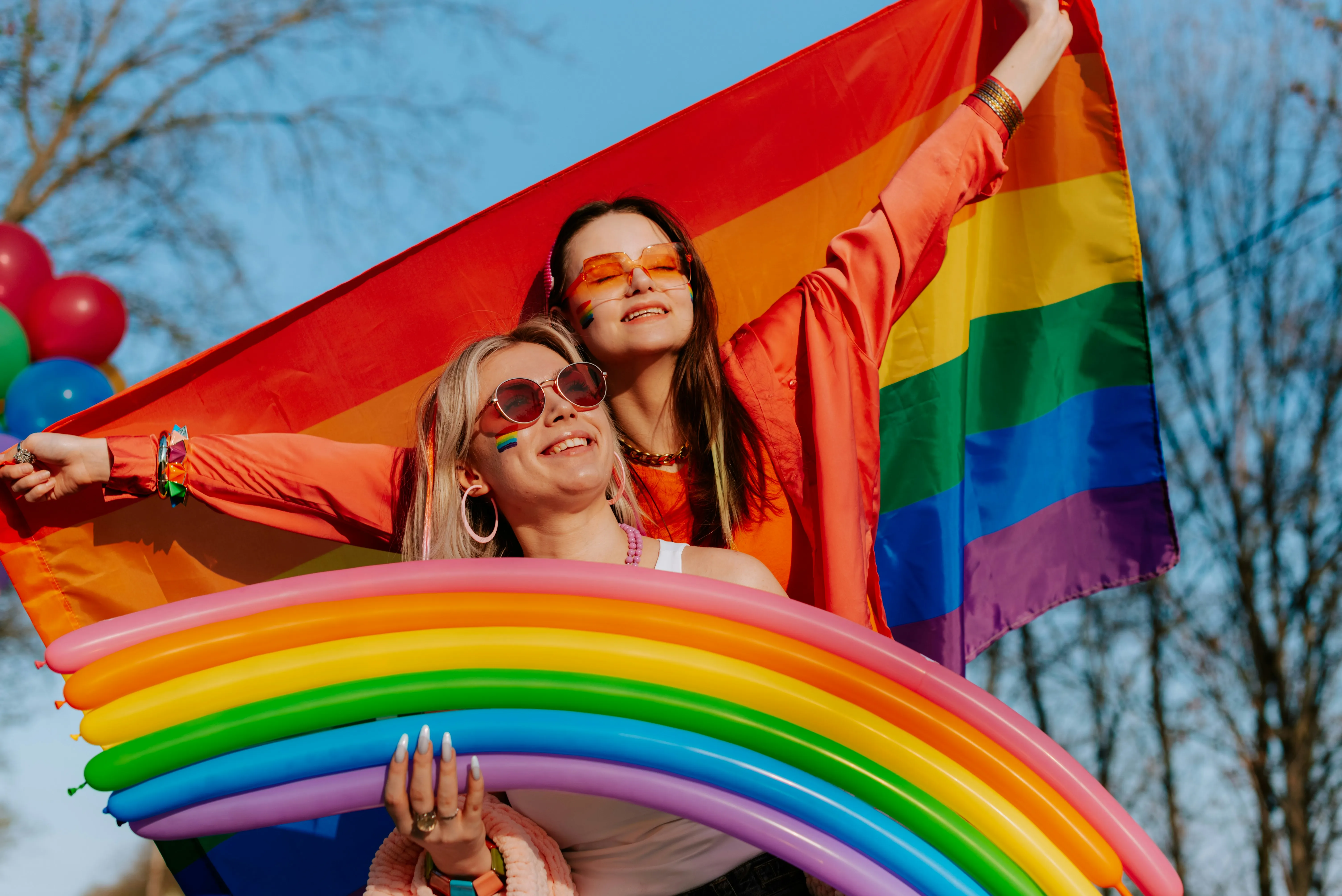 Two happy young women wearing sunglasses, holding a large rainbow pride flag and a rainbow balloon arch, with one having a small rainbow flag painted on her cheek, against a blue sky. Symbolises LGBTQ+ celebrations and inclusive events.