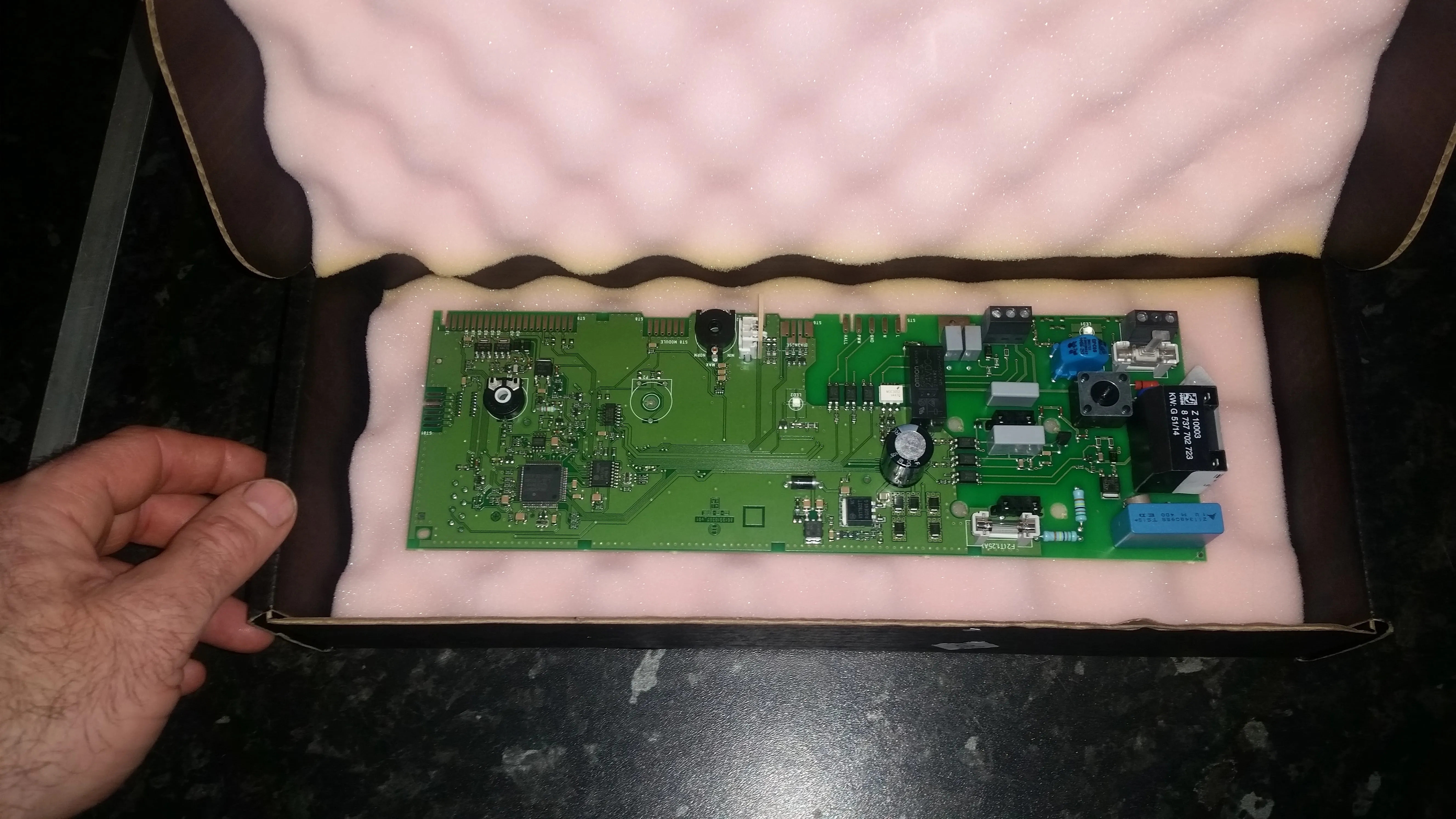 A green circuit board with various electronic components, possibly a boiler PCB, packaged in a box with pink foam.