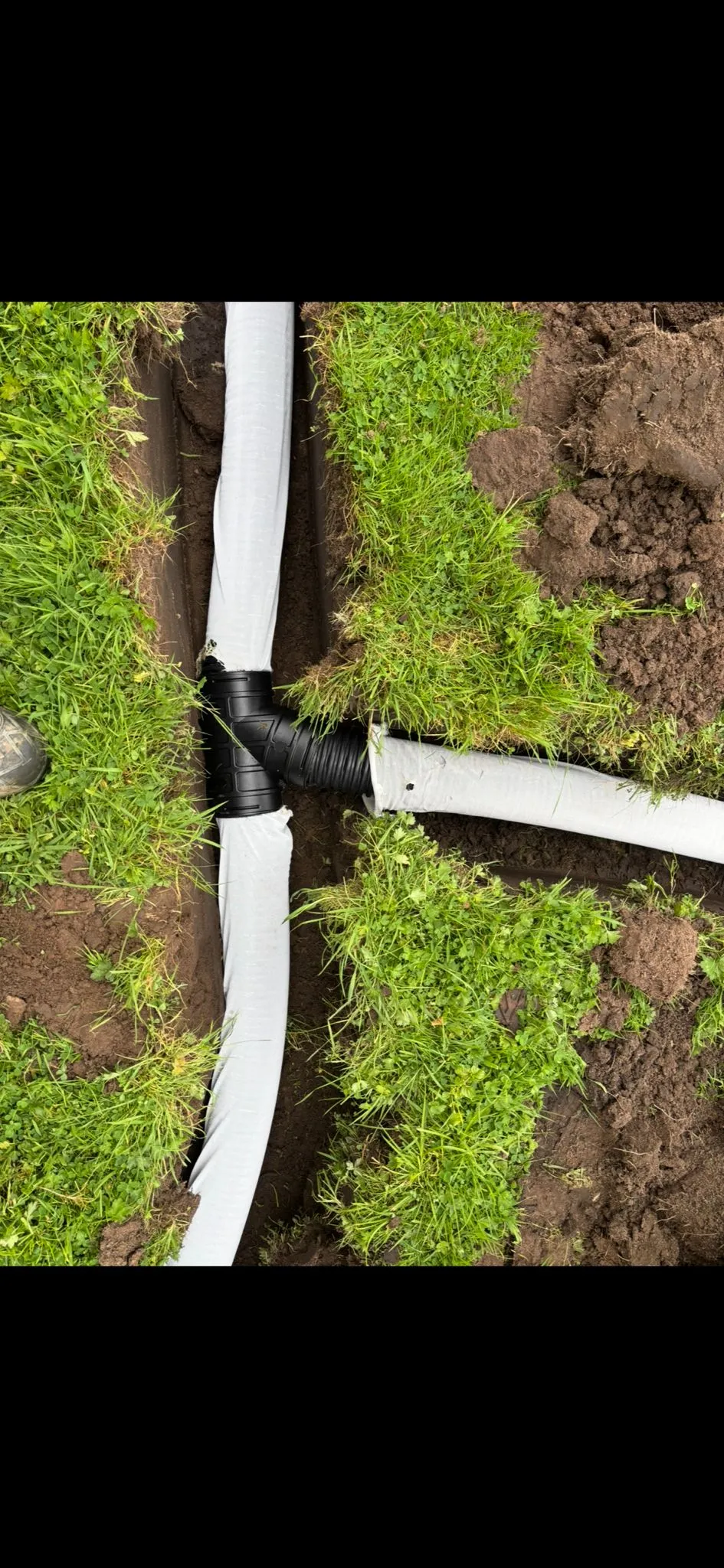Underground drainage pipes being laid in an open trench in a garden, with soil and green grass visible around the excavation.