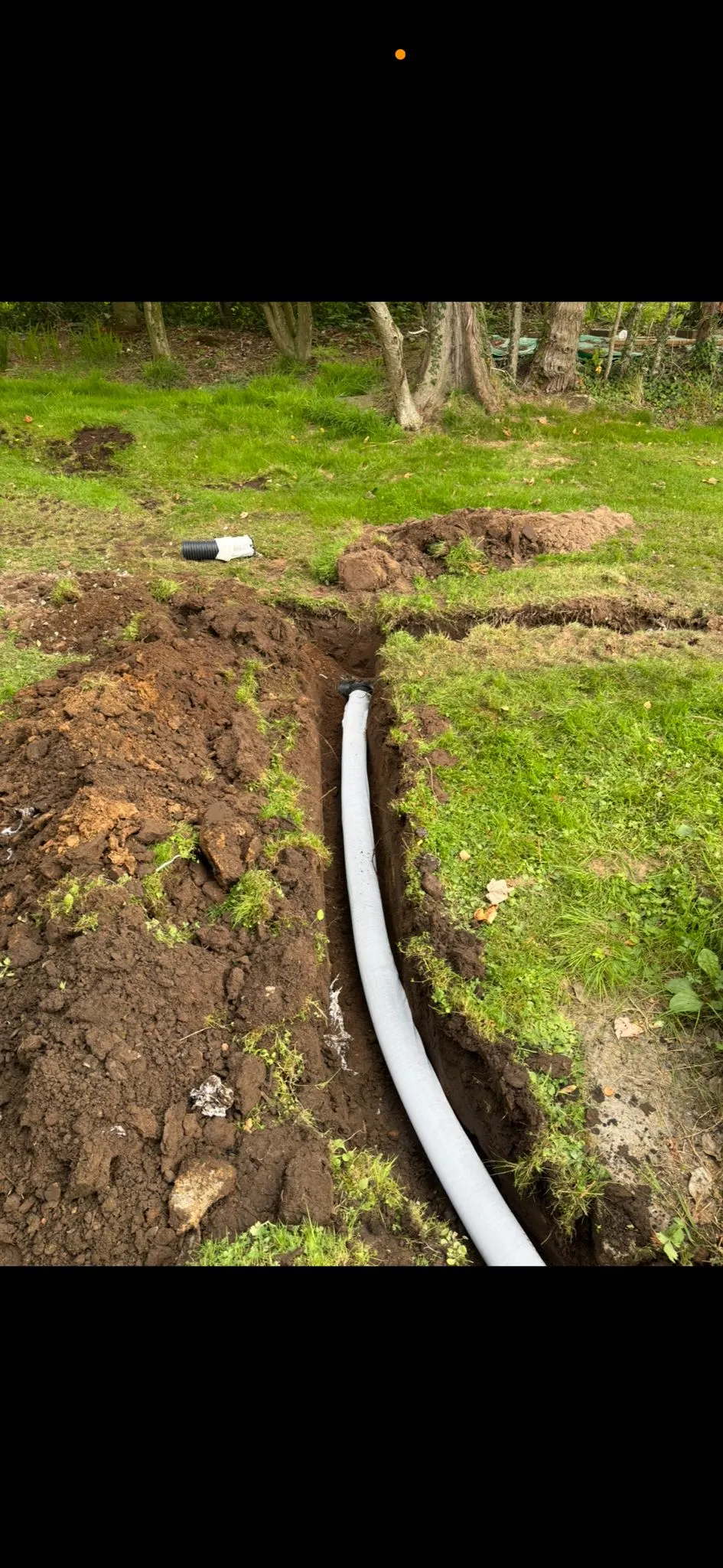 A white drainage pipe laid within an excavated trench in a garden setting, with soil piled on either side.