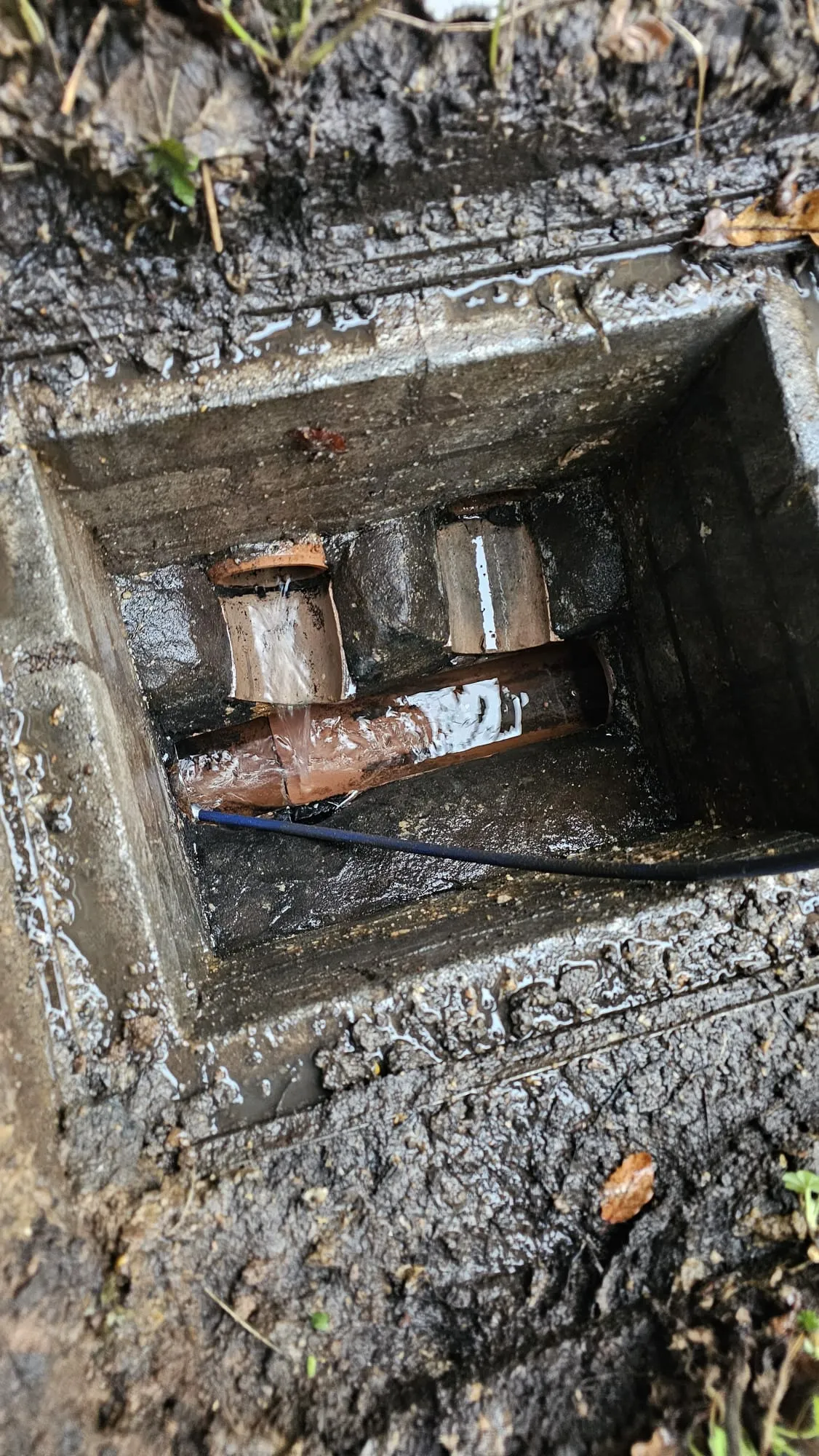 Close-up of a damaged drainage pipe leaking water inside an open inspection chamber, with a blue diagnostic or repair tool visible.