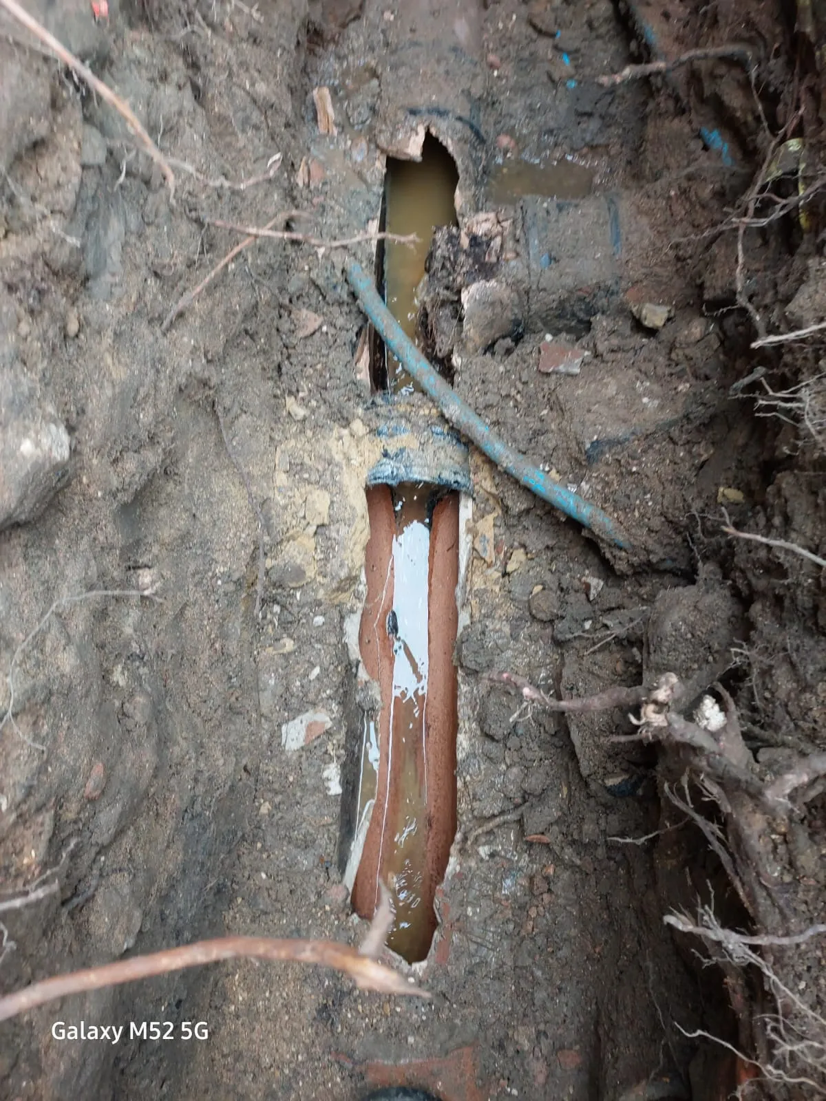 An excavated trench showing a damaged and broken drain pipe with standing water inside, suggesting a drainage repair is needed.