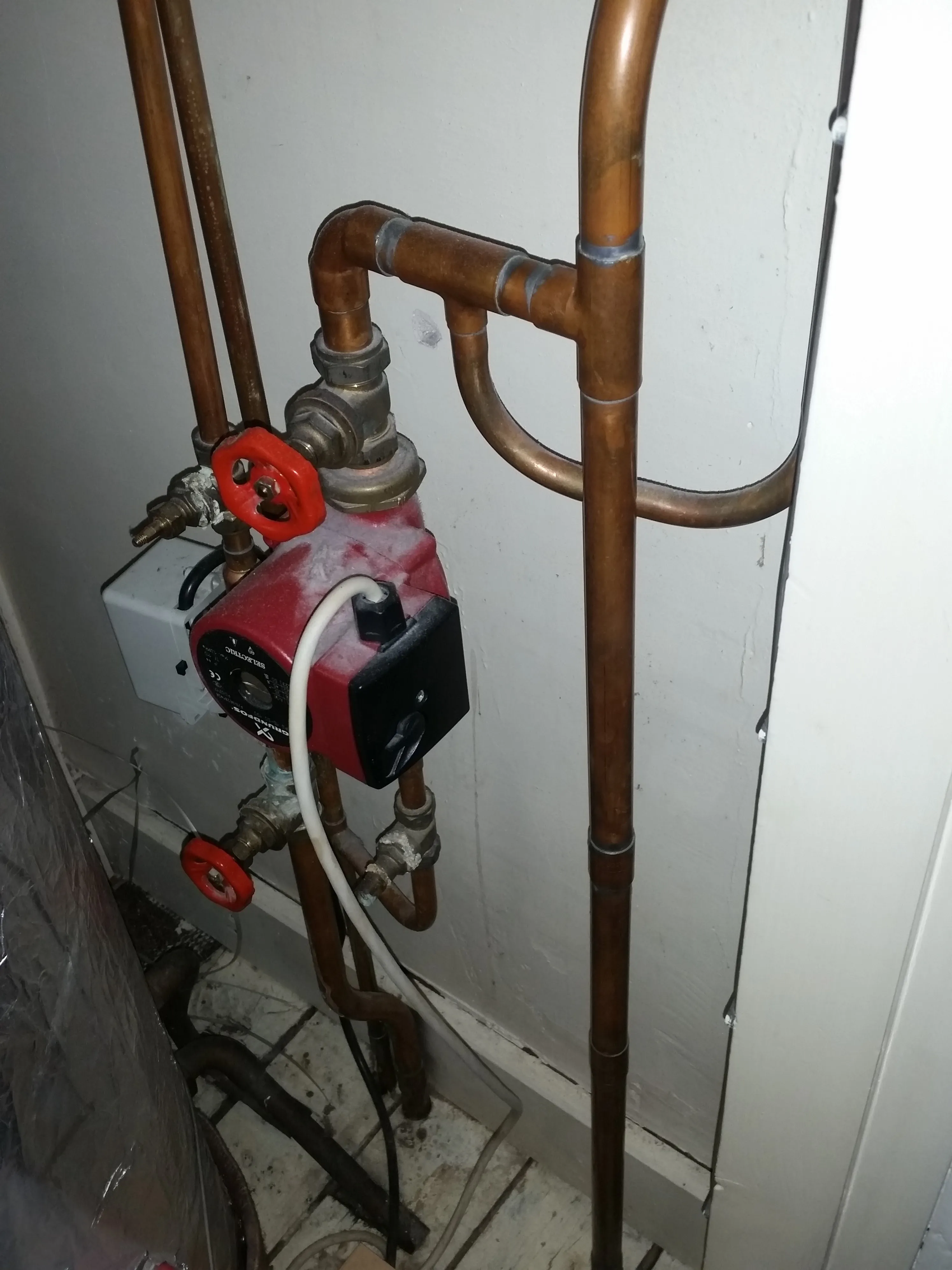 A red central heating pump, likely a Grundfos unit, connected to copper pipes with red isolation valves, against a light-coloured wall.