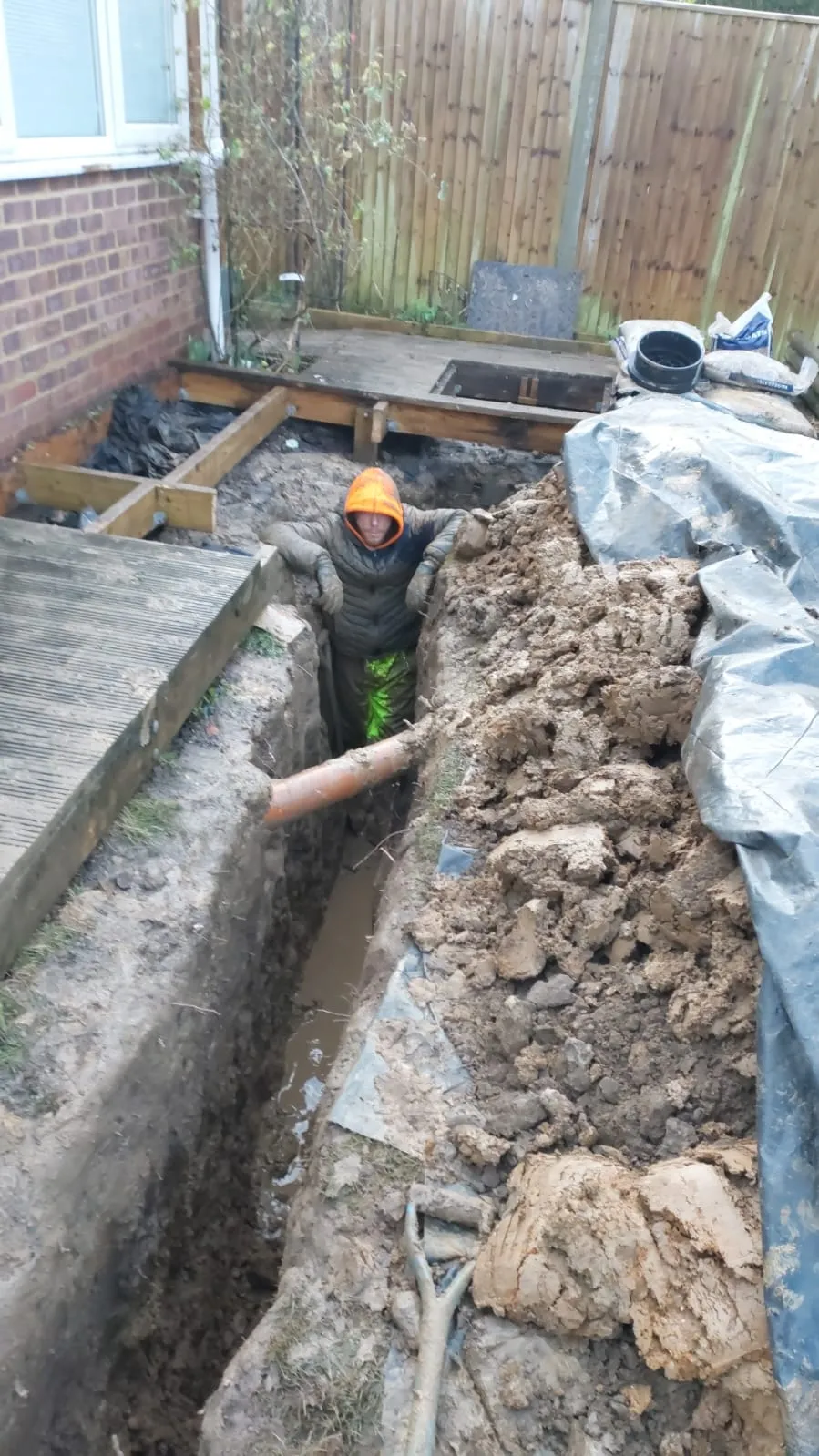 A drainage engineer in an orange hood working in a deep trench filled with muddy water and an exposed drainage pipe, surrounded by excavated soil and a partially dismantled wooden deck, representing drain repair or new installation.