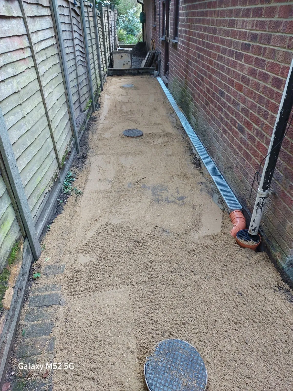 Installation of an ACO channel drain and manhole covers in a residential alleyway, with sandy ground preparation.