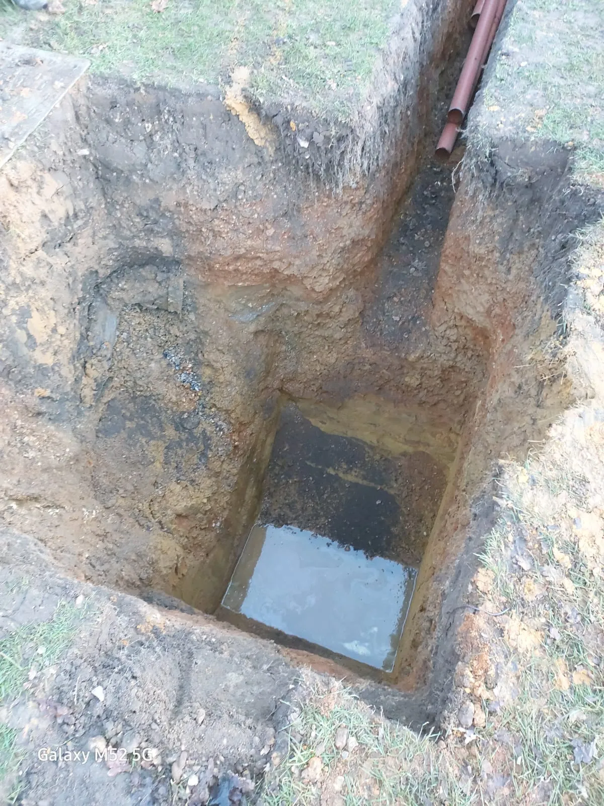 An open trench with brown drainage pipes and accumulated water, indicating ongoing drain repair or new drainage system installation.