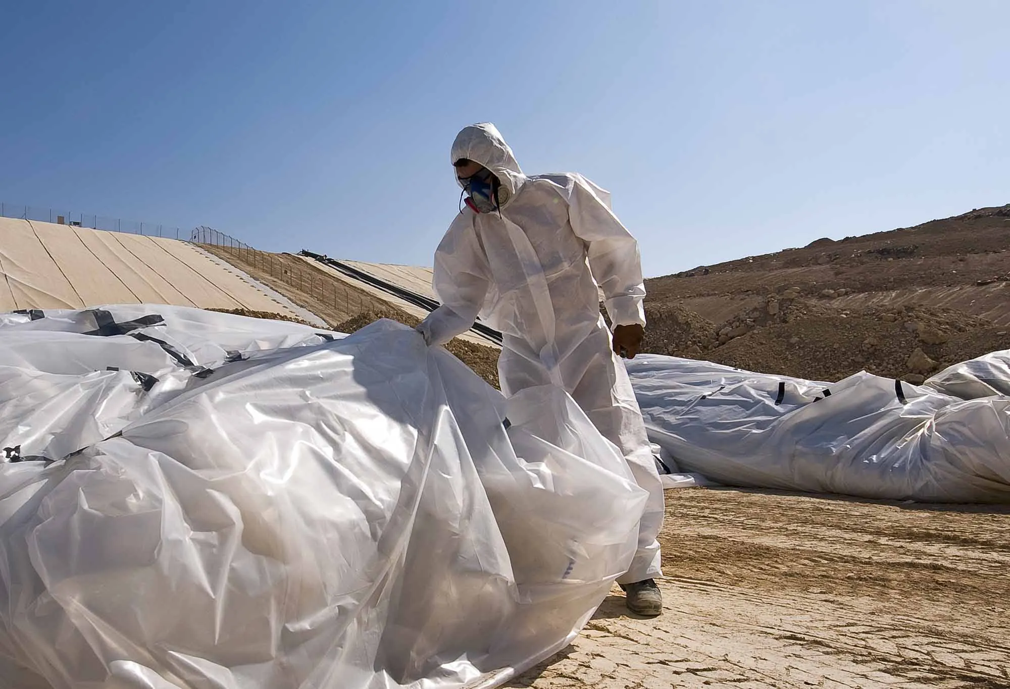 Worker in protective suit and mask handling large white bags of waste outdoors.
