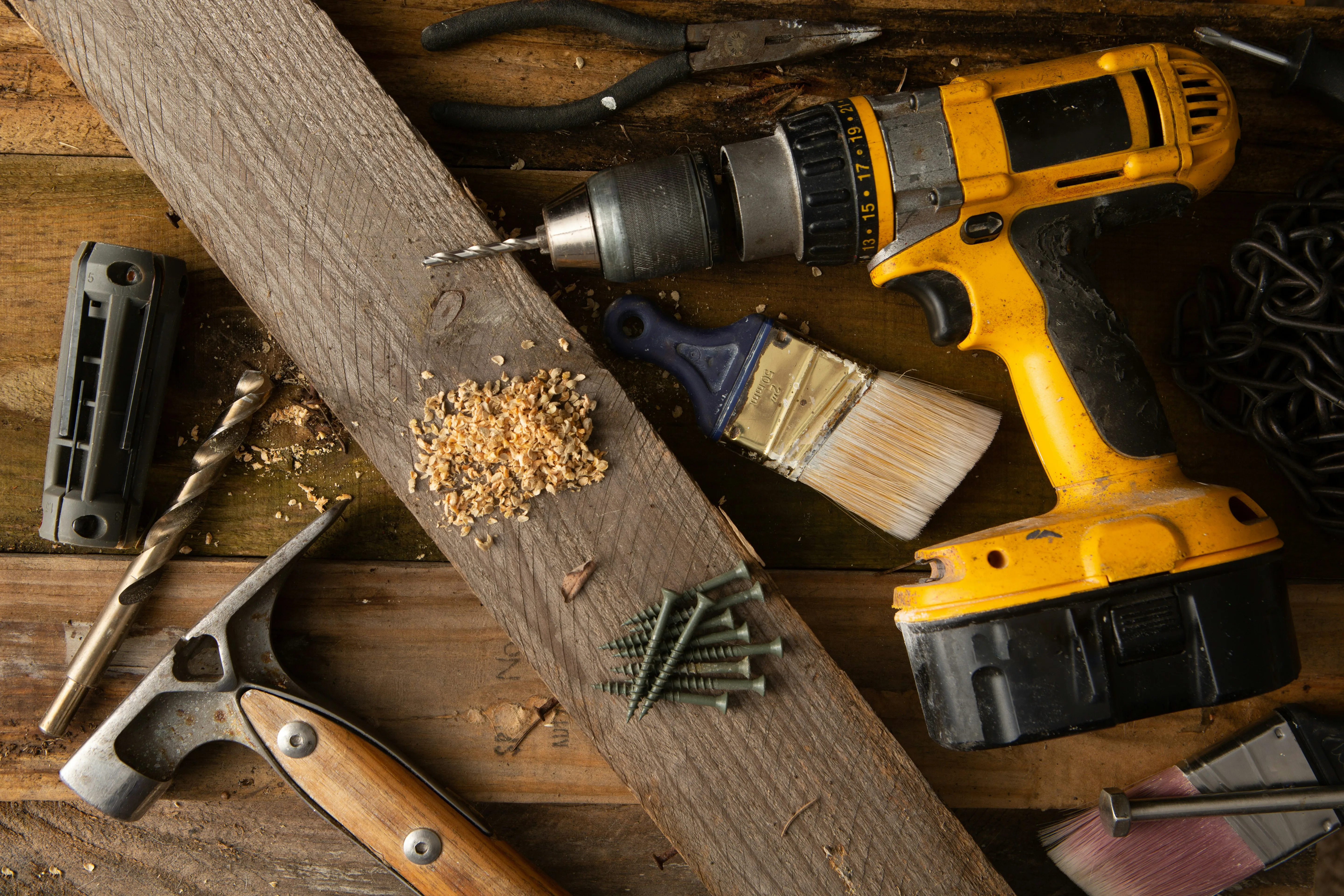 Close-up of roofing tools: a yellow cordless drill, hammer, green screws, and a paintbrush on worn wooden planks, representing expert craftsmanship and ongoing work.
