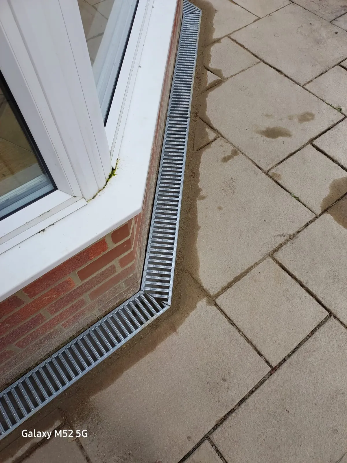 Close-up of a new ACO channel drain with metal grate installed between a brick building wall and paving slabs.